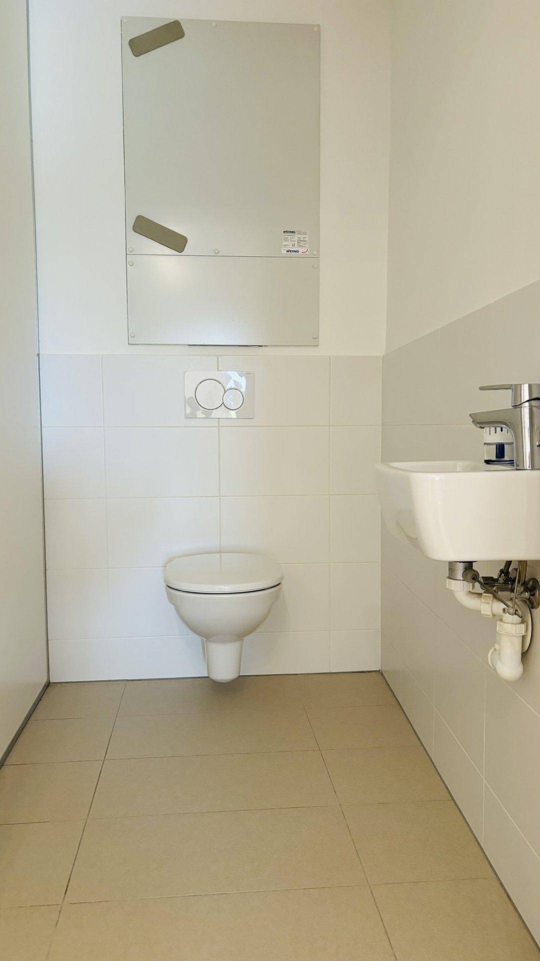 Separates WC mit wandhängender Toilette, kleinem Waschbecken und Fliesenboden.