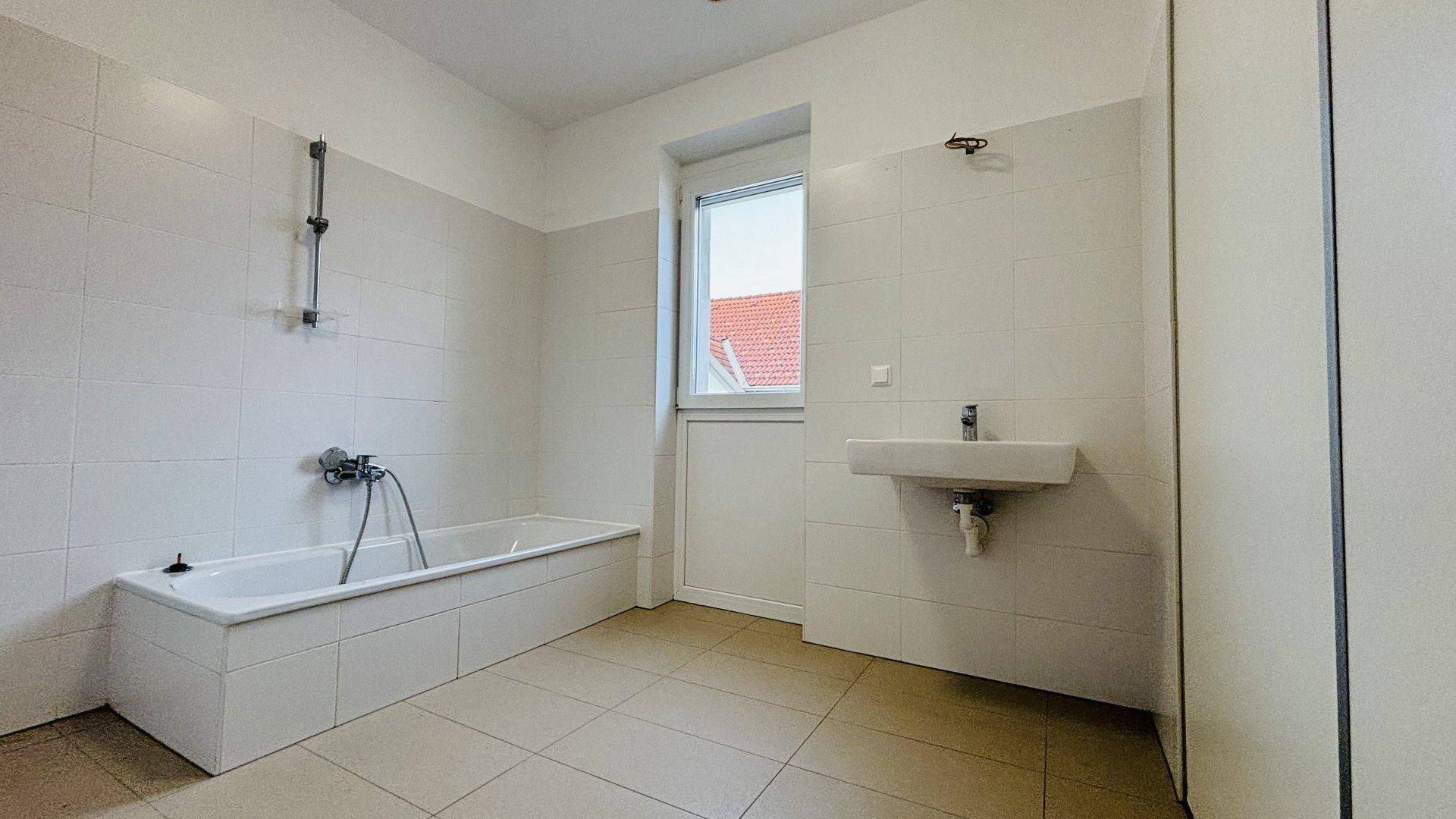 Badezimmer mit Badewanne, Fliesenboden, Waschbecken und Fenster für Tageslicht.