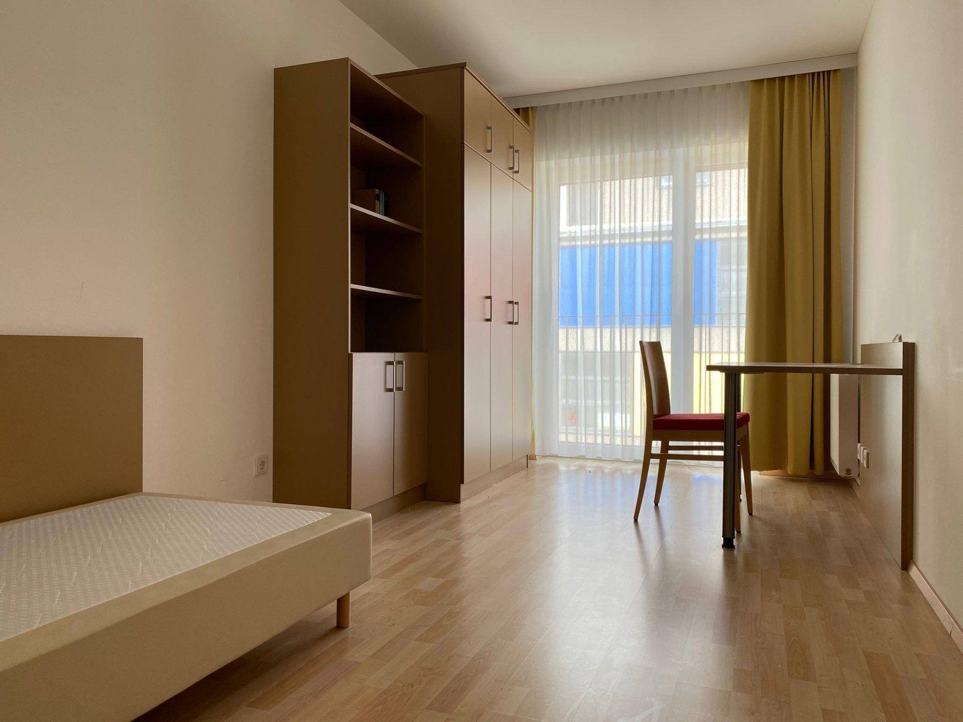 Helles Schlafzimmer mit Einzelbett, Kleiderschrank und Schreibtisch, sowie Zugang zu einem Balkon.