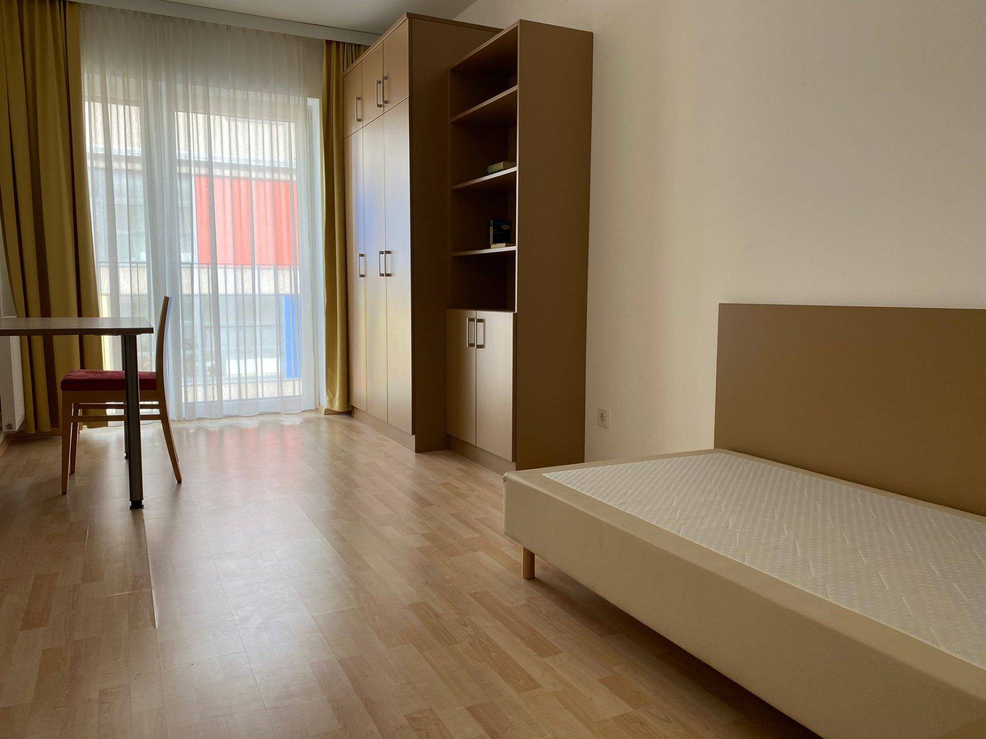 Geräumiges Schlafzimmer mit Laminatboden, Bett, Schrank und Schreibtisch, hell durch große Fenster.