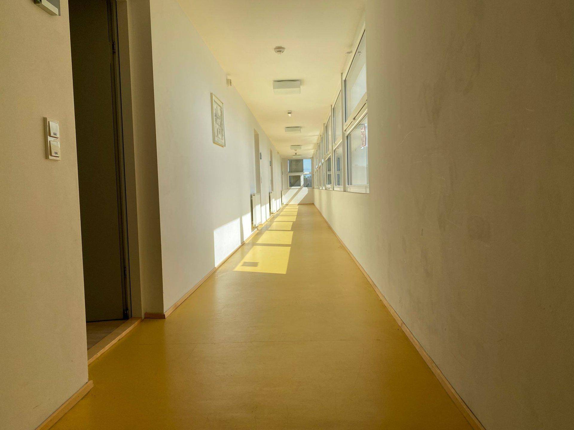 Langer, heller Flur mit gelbem Boden und Fenstern, die viel Tageslicht hereinlassen.