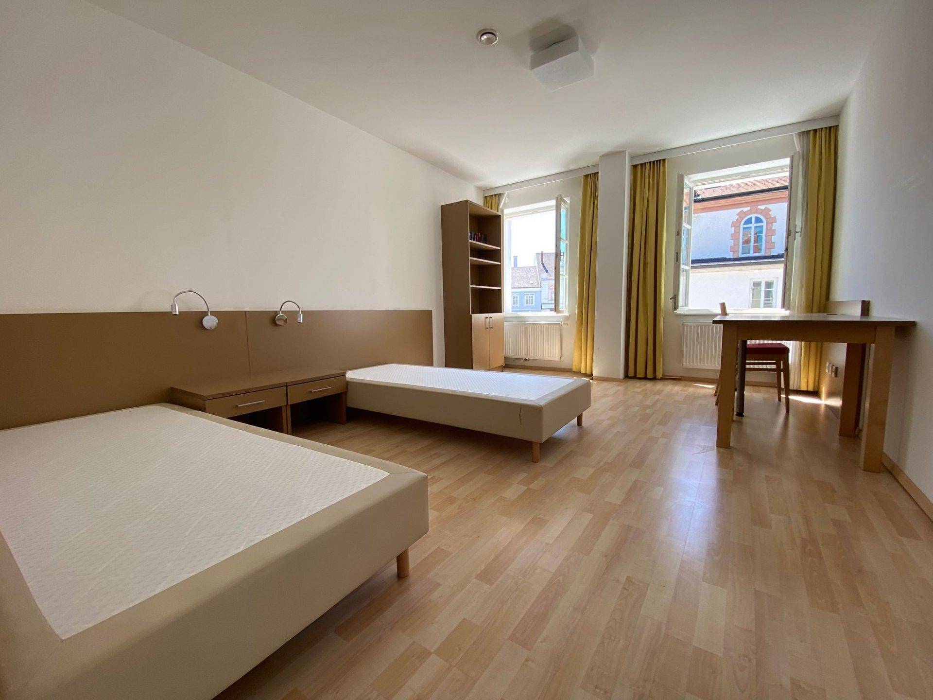Helles Schlafzimmer mit zwei Einzelbetten, Schreibtisch und gelben Vorhängen an den Fenstern.