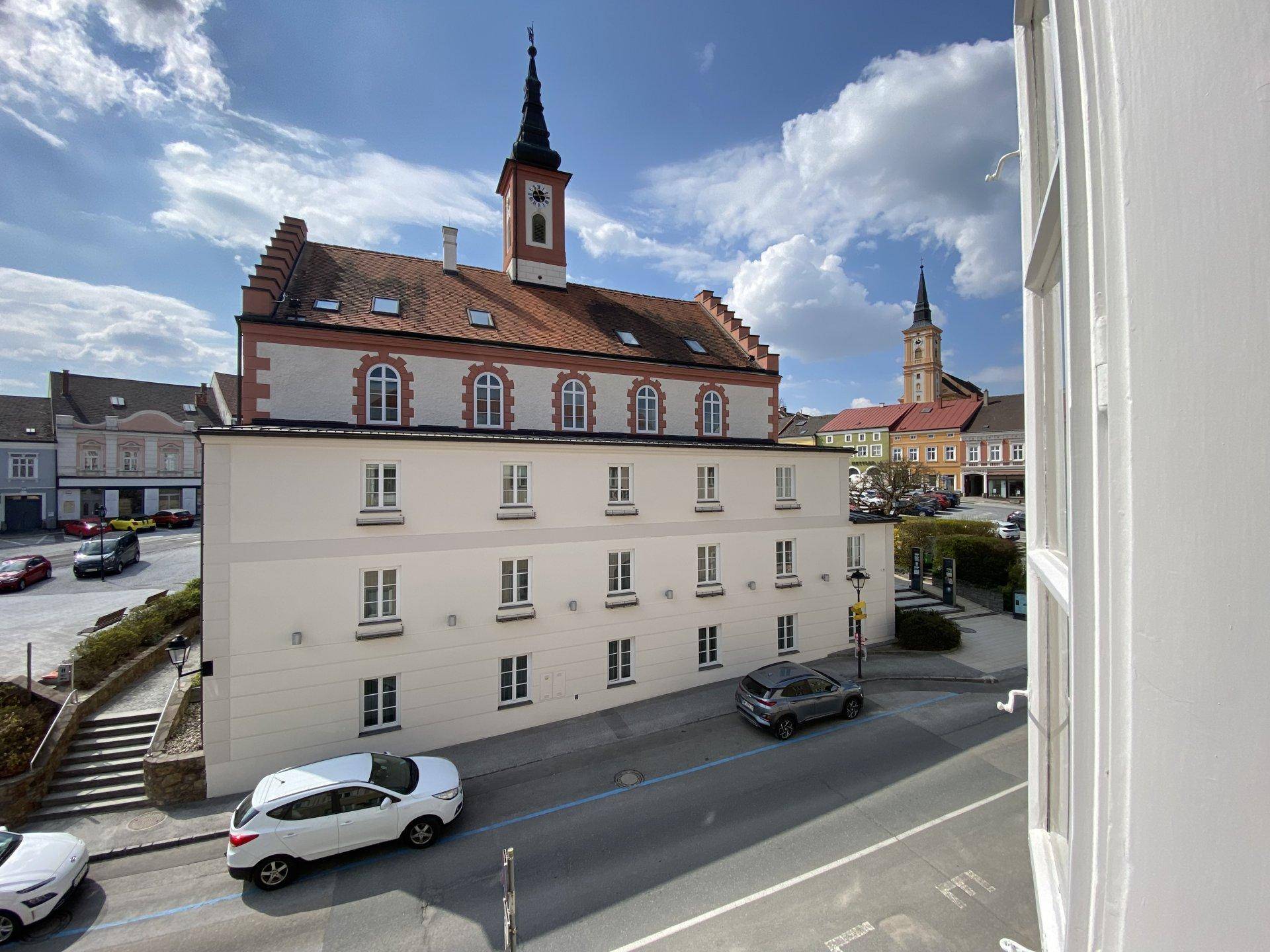 Malerischer Blick auf einen historischen Stadtplatz mit Rathaus und weiteren Gebäuden unter blauem Himmel.