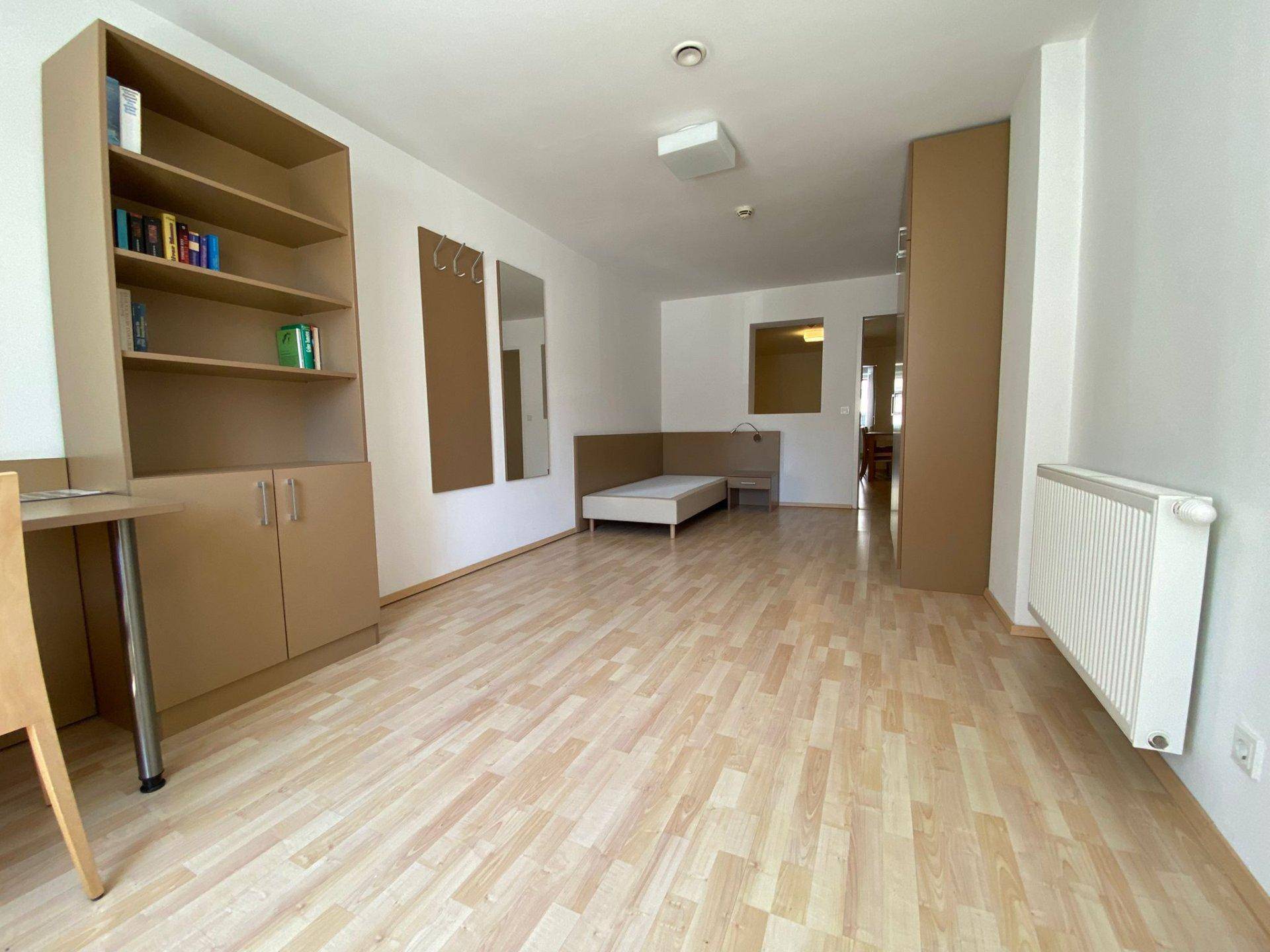 Geräumiger Wohnbereich mit Laminatboden, Regal und einem Bett in einem Studio-Apartment.