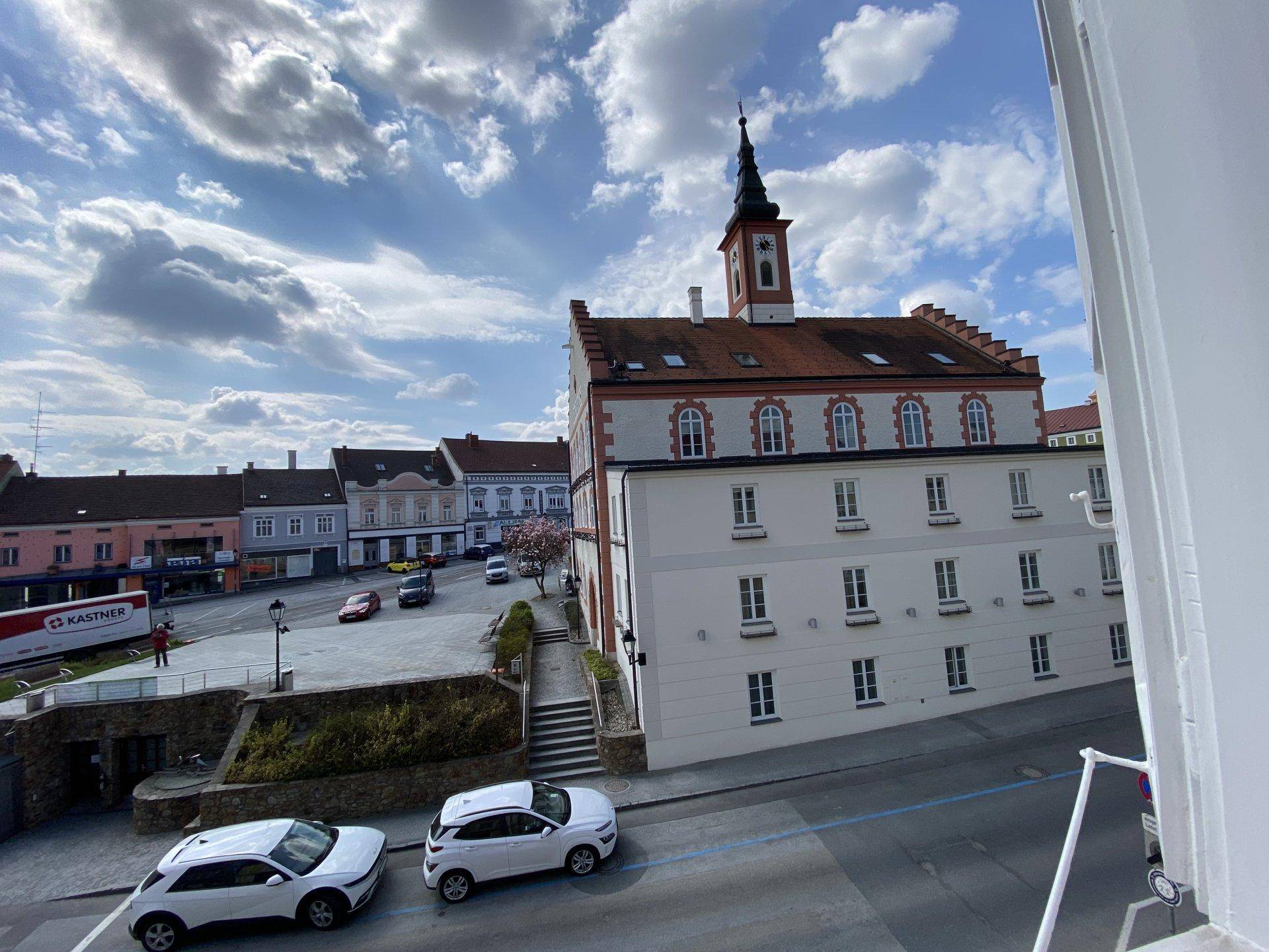 Blick auf einen belebten Stadtplatz mit historischen Gebäuden und parkenden Autos.
