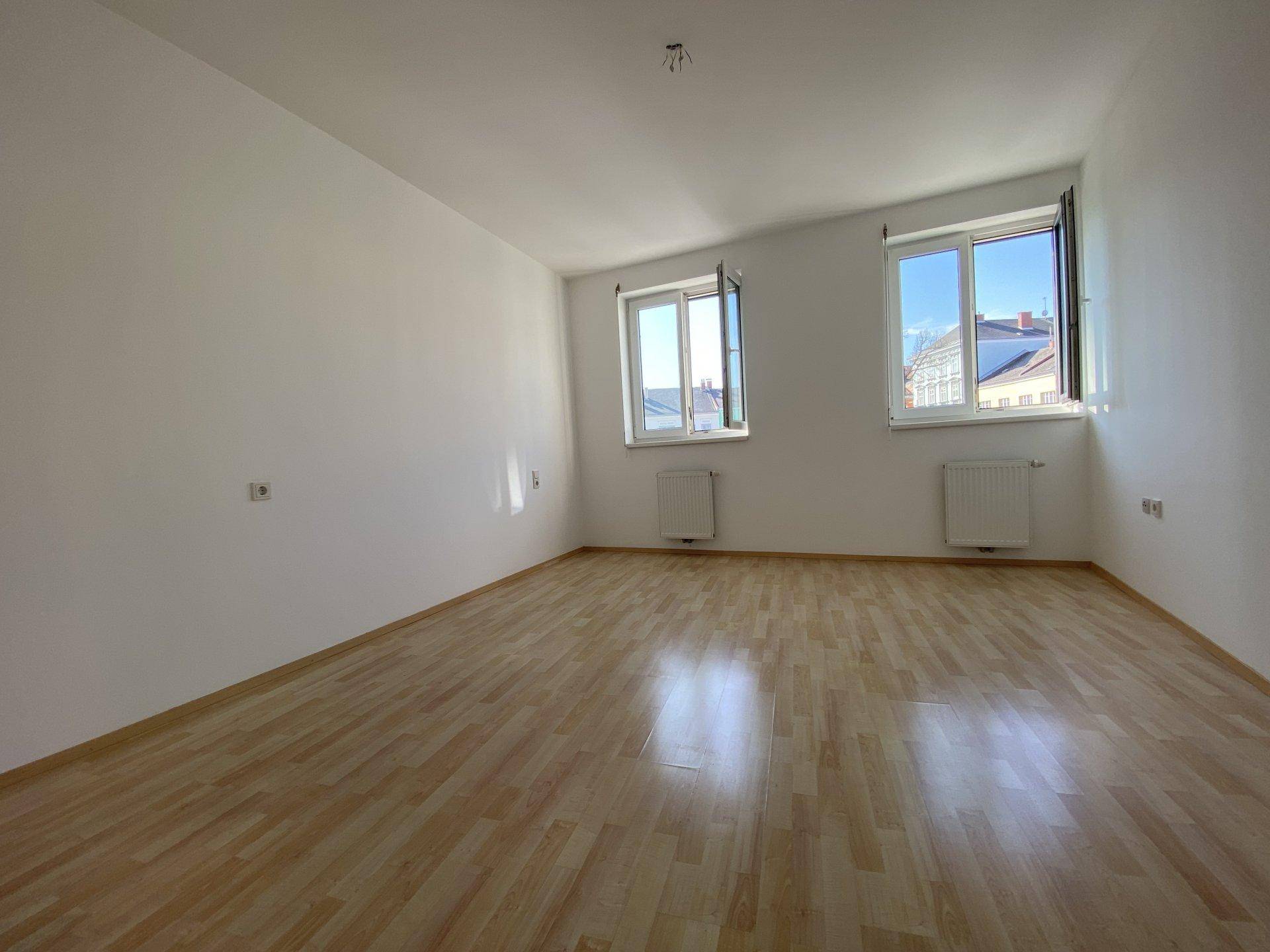 Geräumiger, heller Raum mit großen Fenstern und hellem Laminatboden.