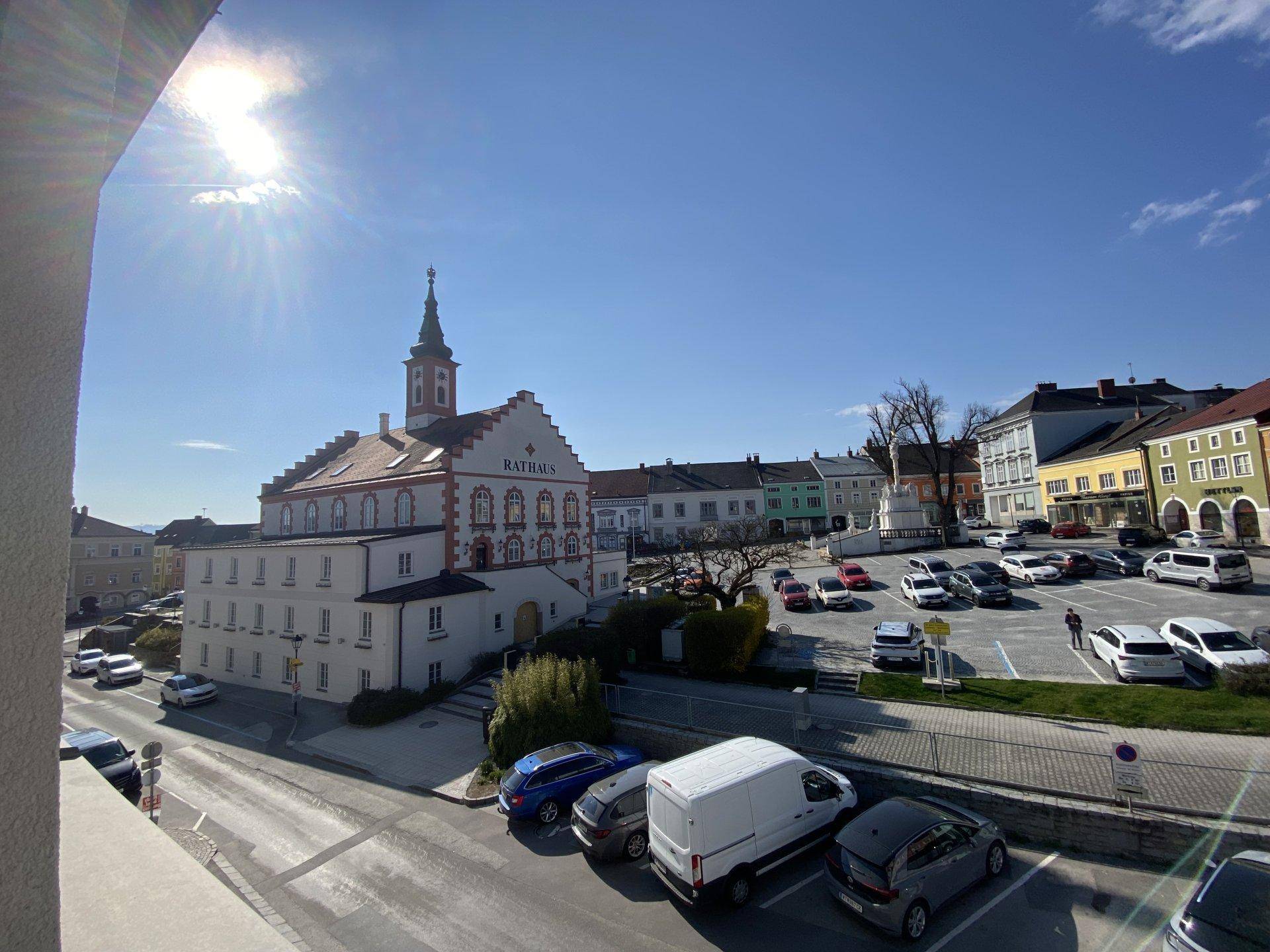 Blick auf den belebten Stadtplatz mit historischem Rathaus und parkenden Autos.