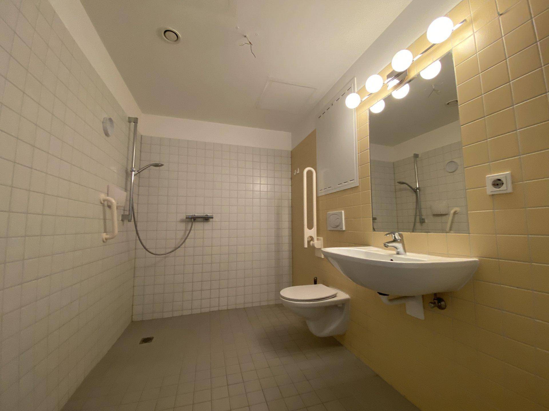 Barrierefreies Badezimmer mit ebenerdiger Dusche, Haltegriffen und gelben Fliesen.