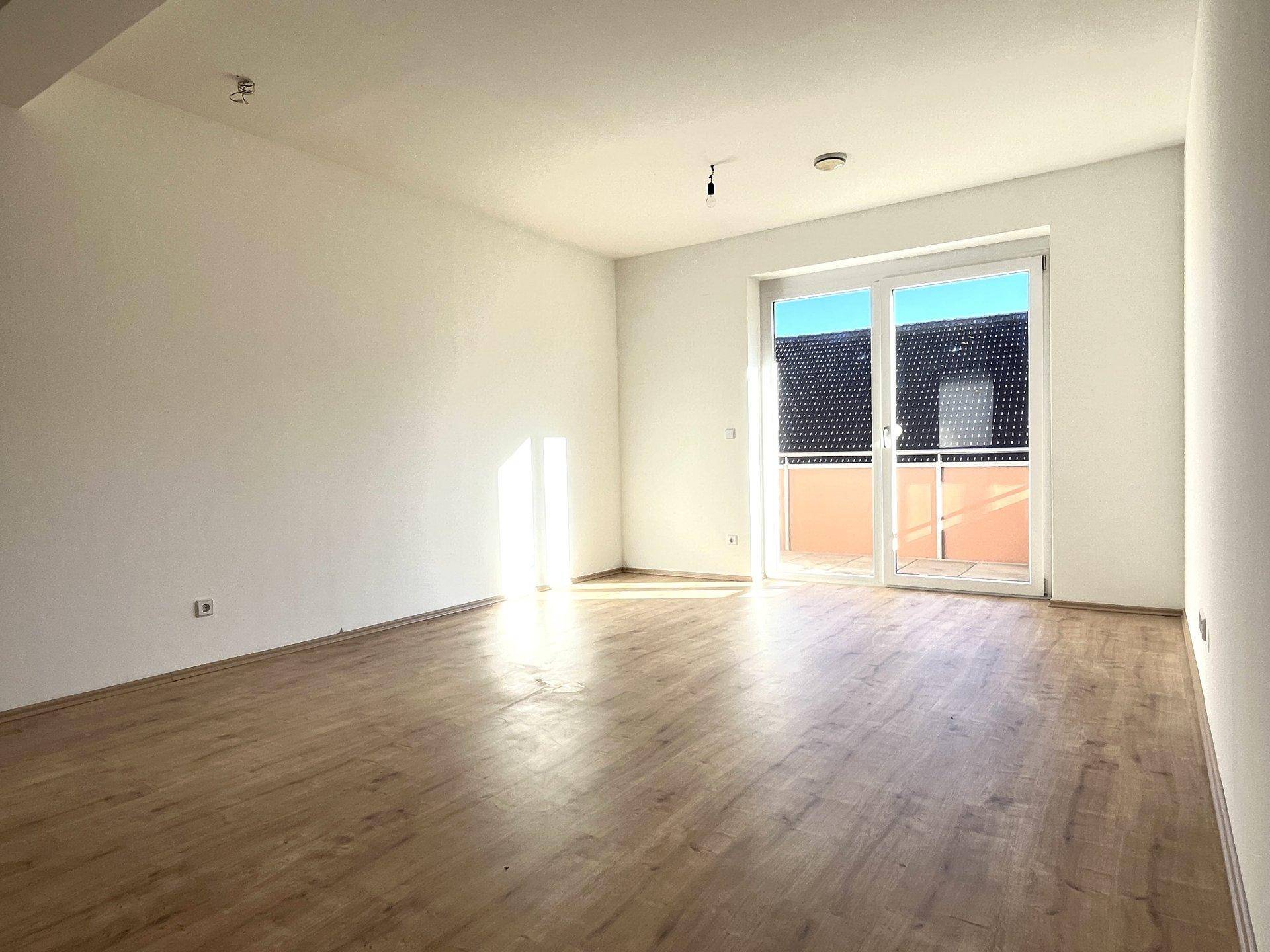 Lichtdurchflutetes Wohnzimmer mit Holzboden und großem Fenster, das Zugang zu einem Balkon bietet.