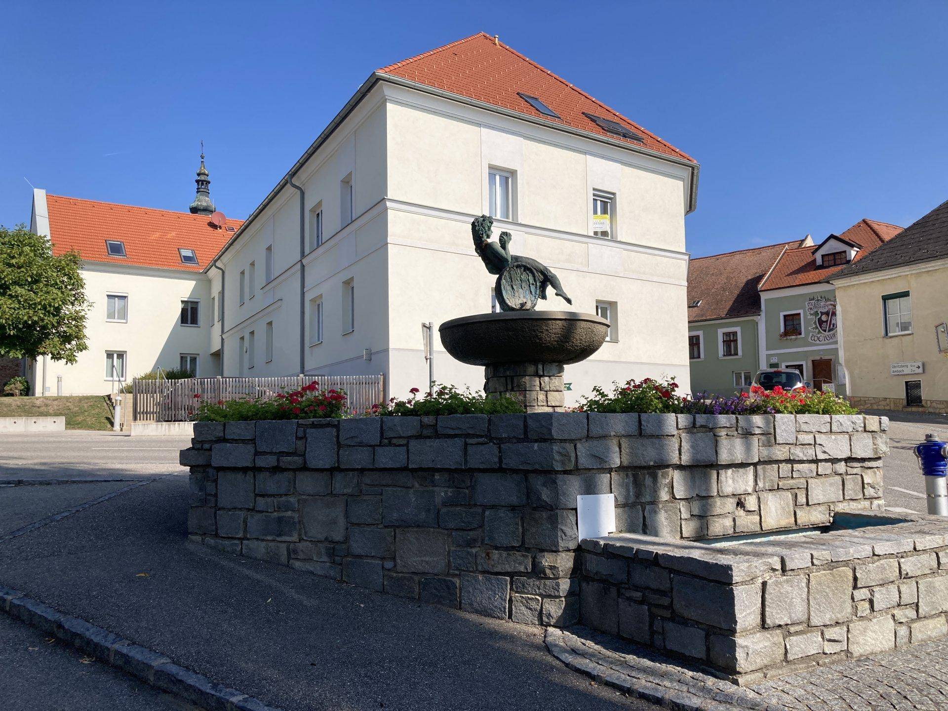 Historisches Gebäude mit rotem Ziegeldach und einem dekorativen Steinbrunnen auf dem Vorplatz.