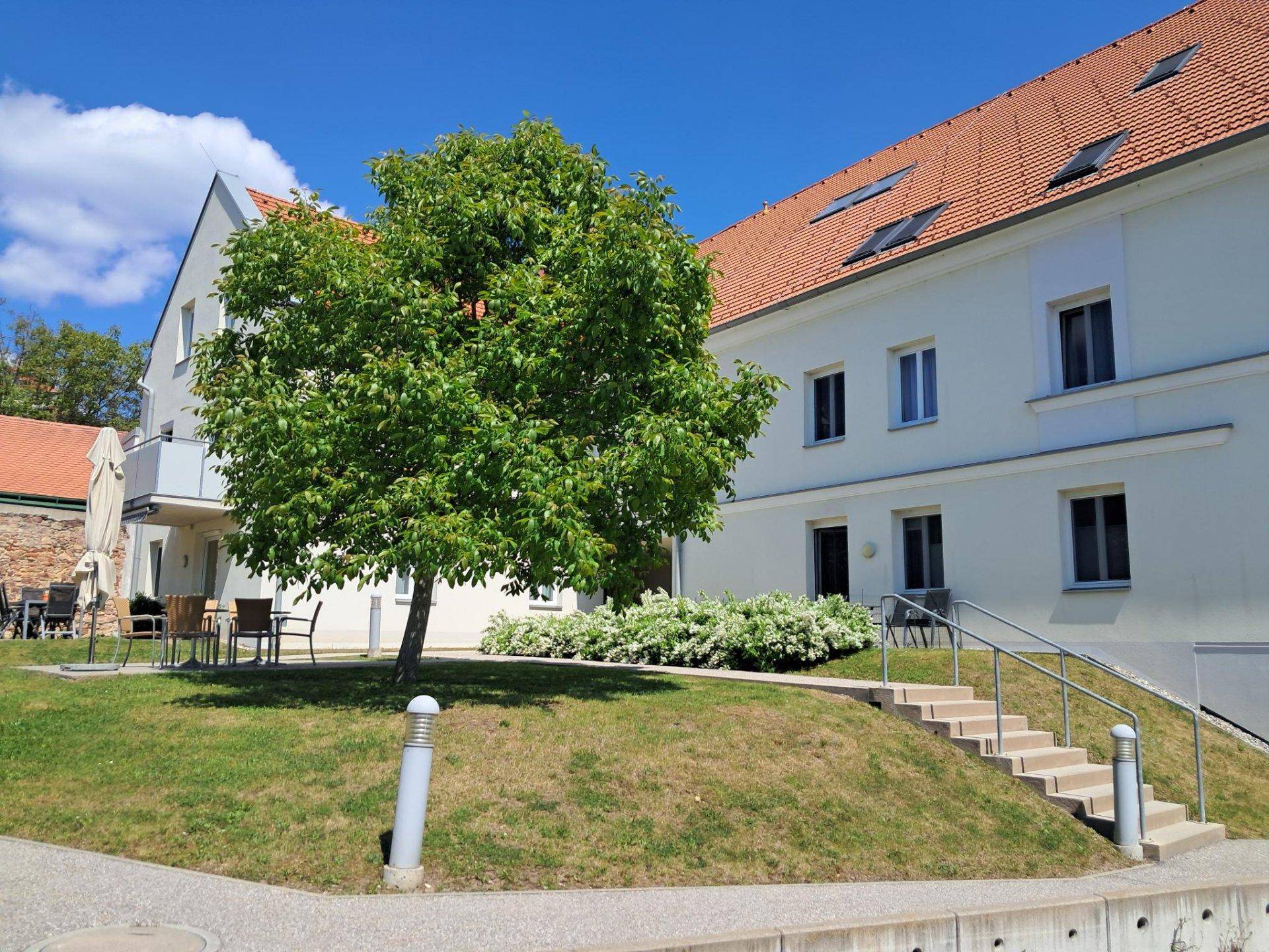 Mehrfamilienhaus mit rotem Ziegeldach, Außentreppe und einer kleinen Terrasse im Gemeinschaftsbereich.