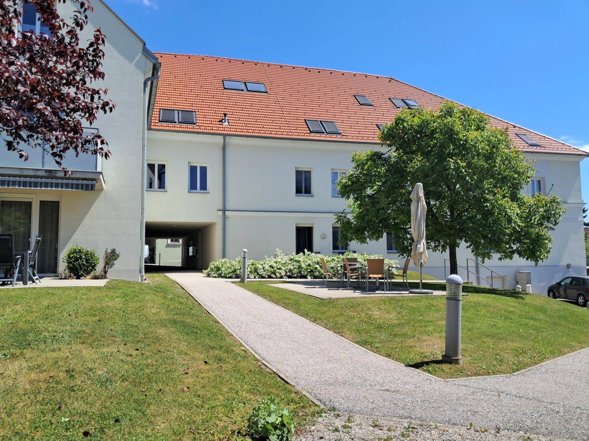 Weißes Mehrfamilienhaus mit rotem Ziegeldach, umgeben von einer gepflegten Grünfläche und Bäumen.