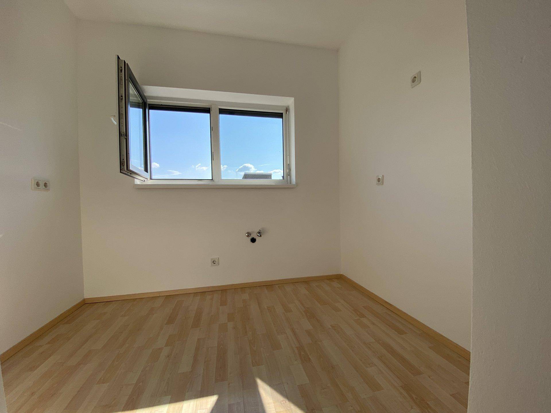 Leerer Raum mit hellem Laminatboden, Fenster und Anschlüssen an der Wand.