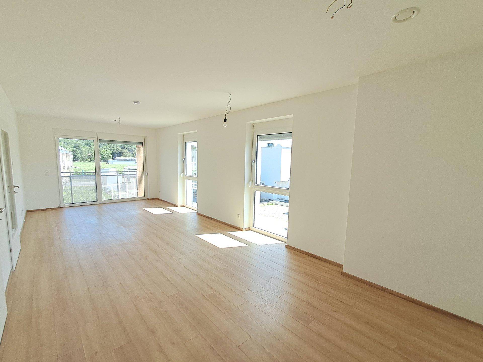 Lichtdurchflutetes Wohnzimmer mit hellem Laminatboden und großen Fenstern, die viel Tageslicht hereinlassen.
