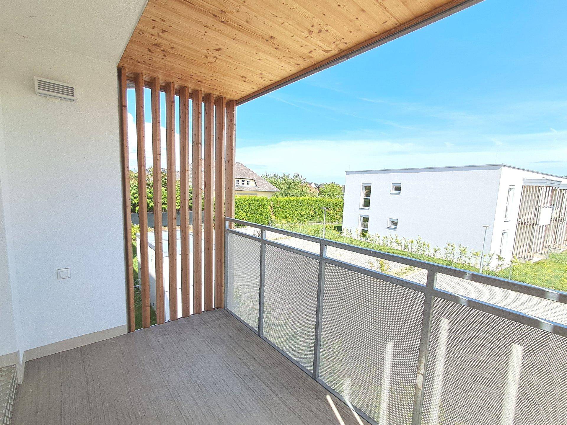 Heller Balkon mit Holzelementen und einem weiten Blick über die grüne Umgebung und weitere Gebäude.