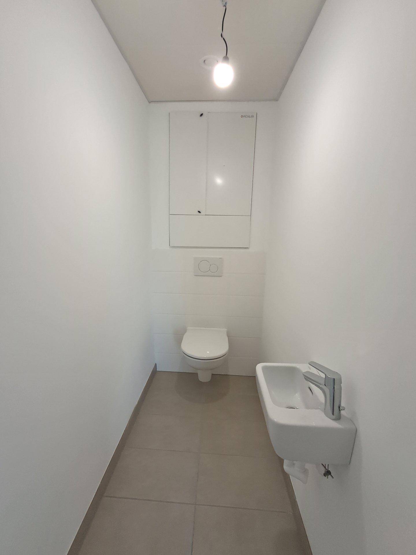 Separates WC mit wandhängender Toilette, kleinem Waschbecken und hellen Fliesen.