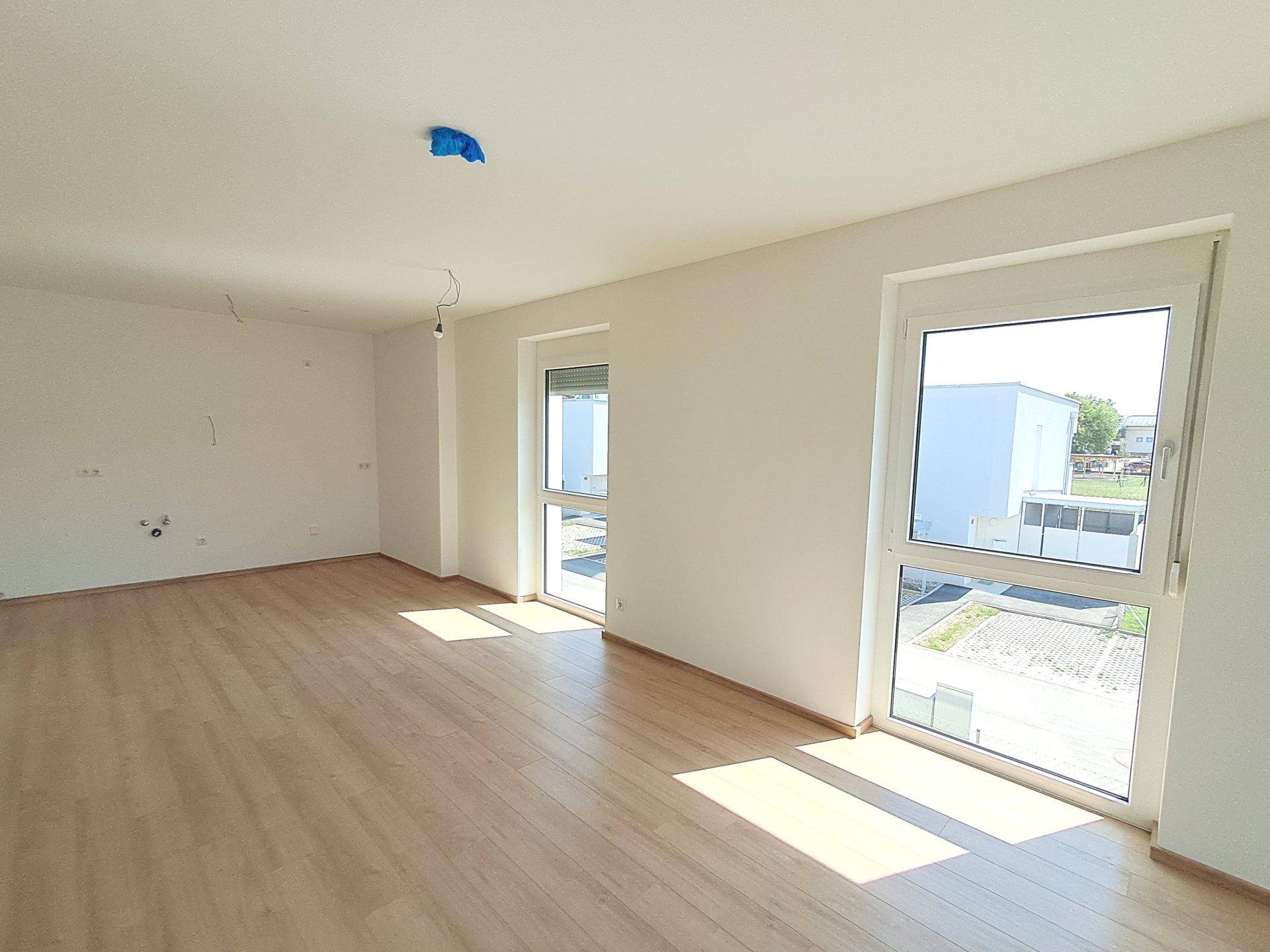 Geräumiges Wohnzimmer mit hellem Laminatboden und großen Fenstern, die viel Tageslicht hereinlassen.