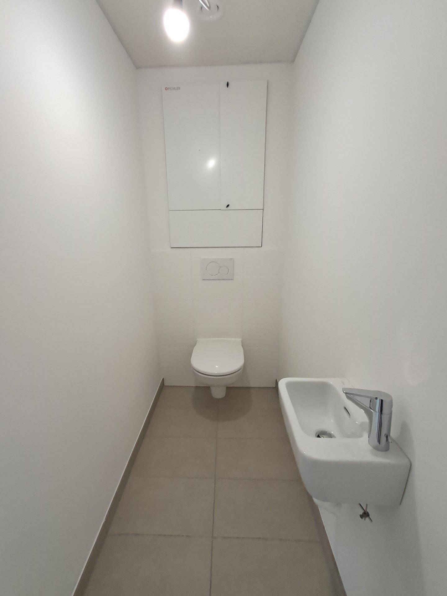 Separates WC mit wandhängender Toilette, kleinem Waschbecken und hellen Fliesen an Wänden und Boden.
