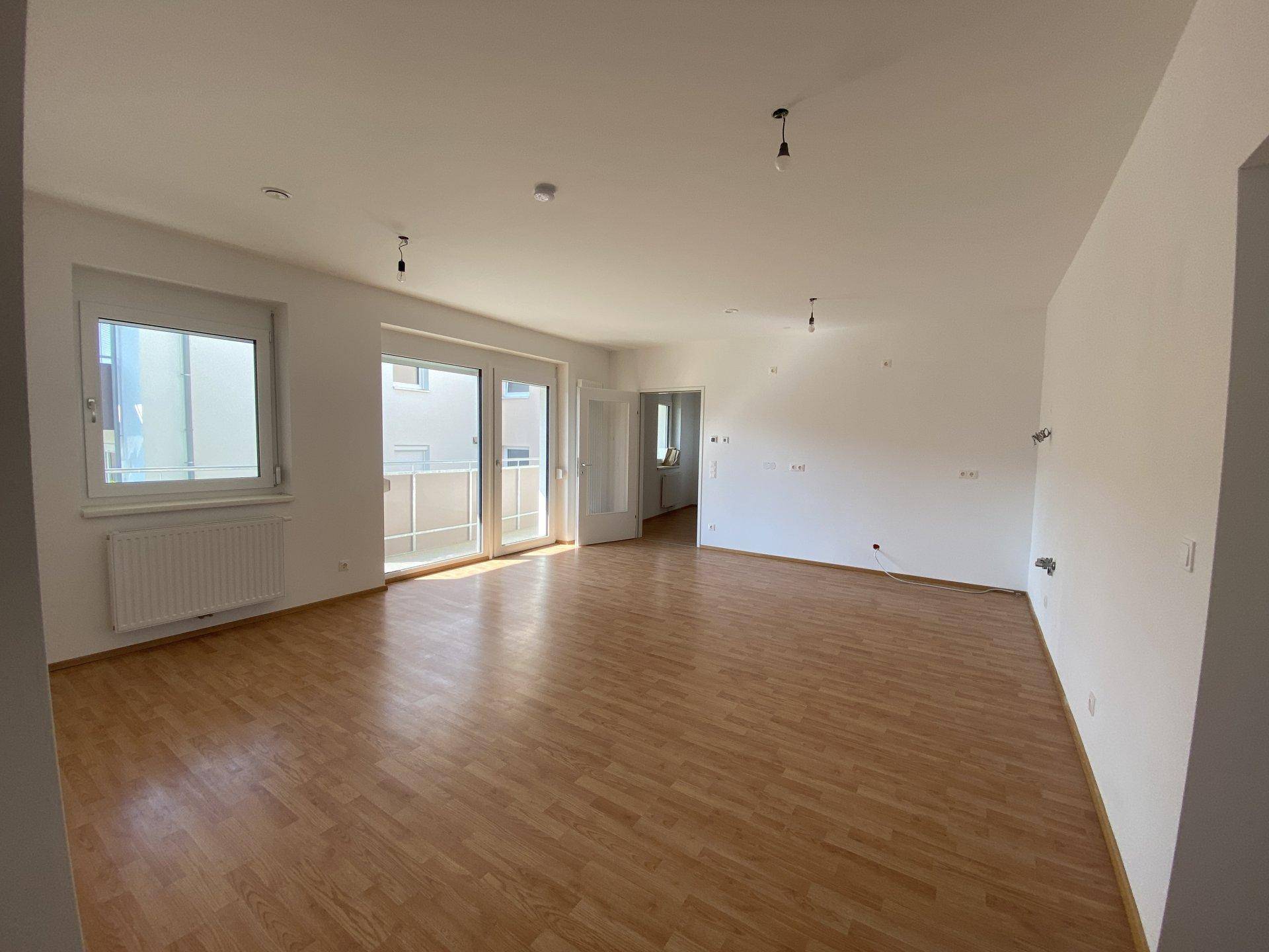 Helles, leeres Wohnzimmer mit Parkettboden, großen Fenstern und Zugang zum Balkon.