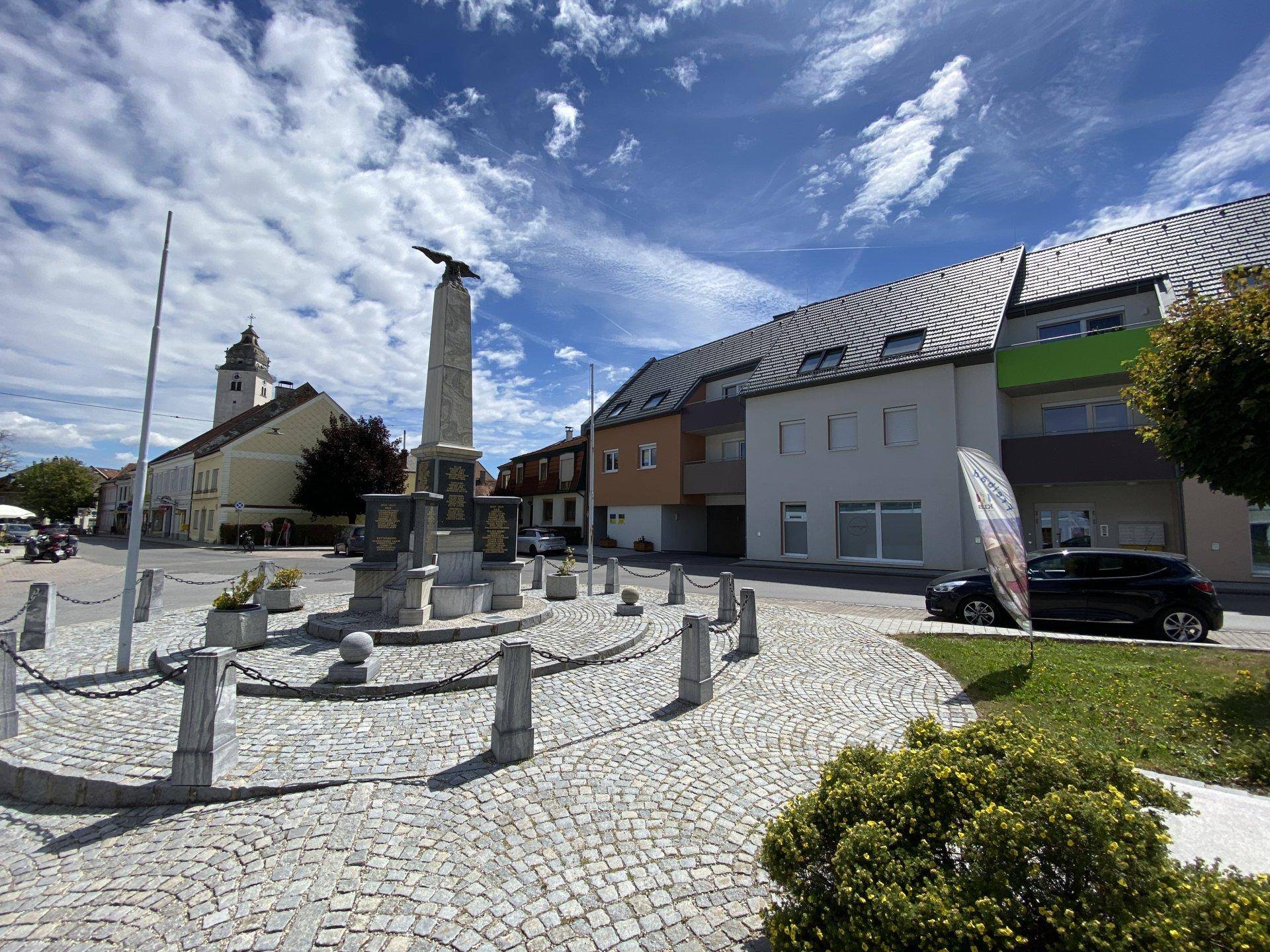 Ansicht der Wohngebäude an einem belebten Platz mit Kopfsteinpflaster und einem Denkmal.