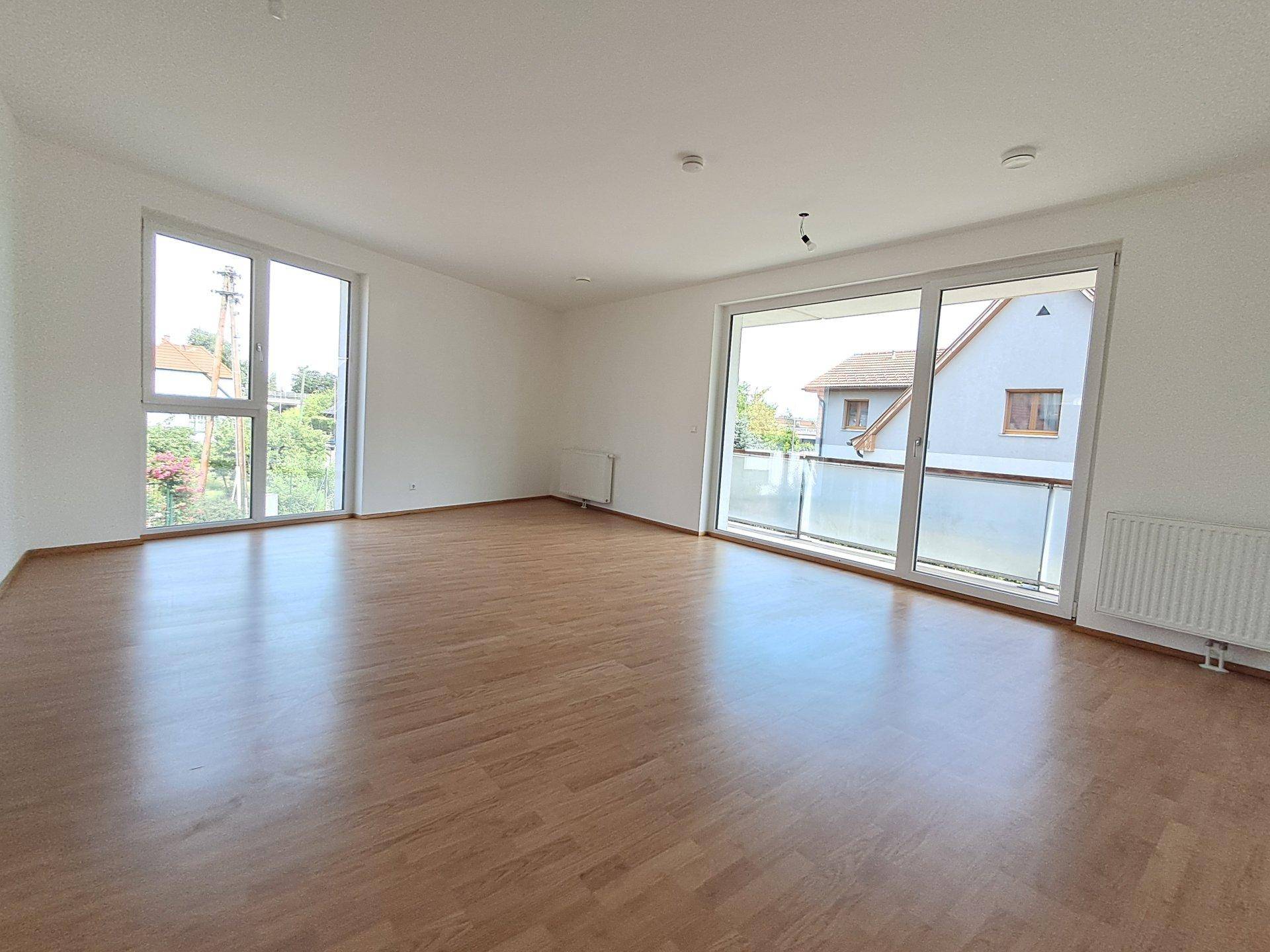 Helles, leeres Wohnzimmer mit Laminatboden, großen Fenstern und Zugang zum Balkon.