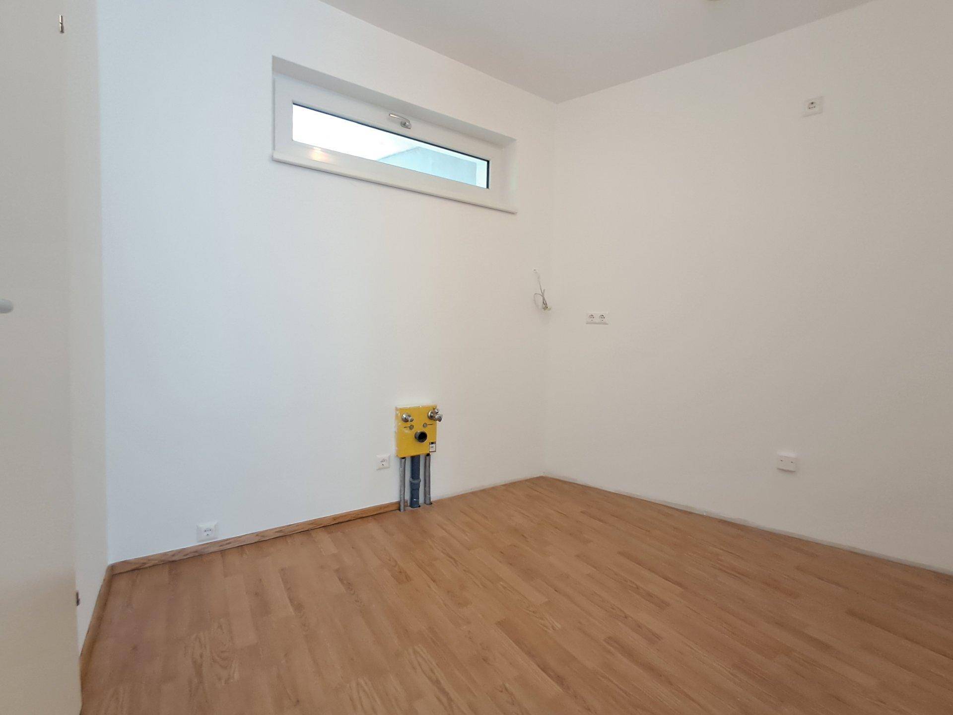 Heller Hauswirtschaftsraum mit Laminatboden, Fenster und Anschlüssen für Waschmaschine.