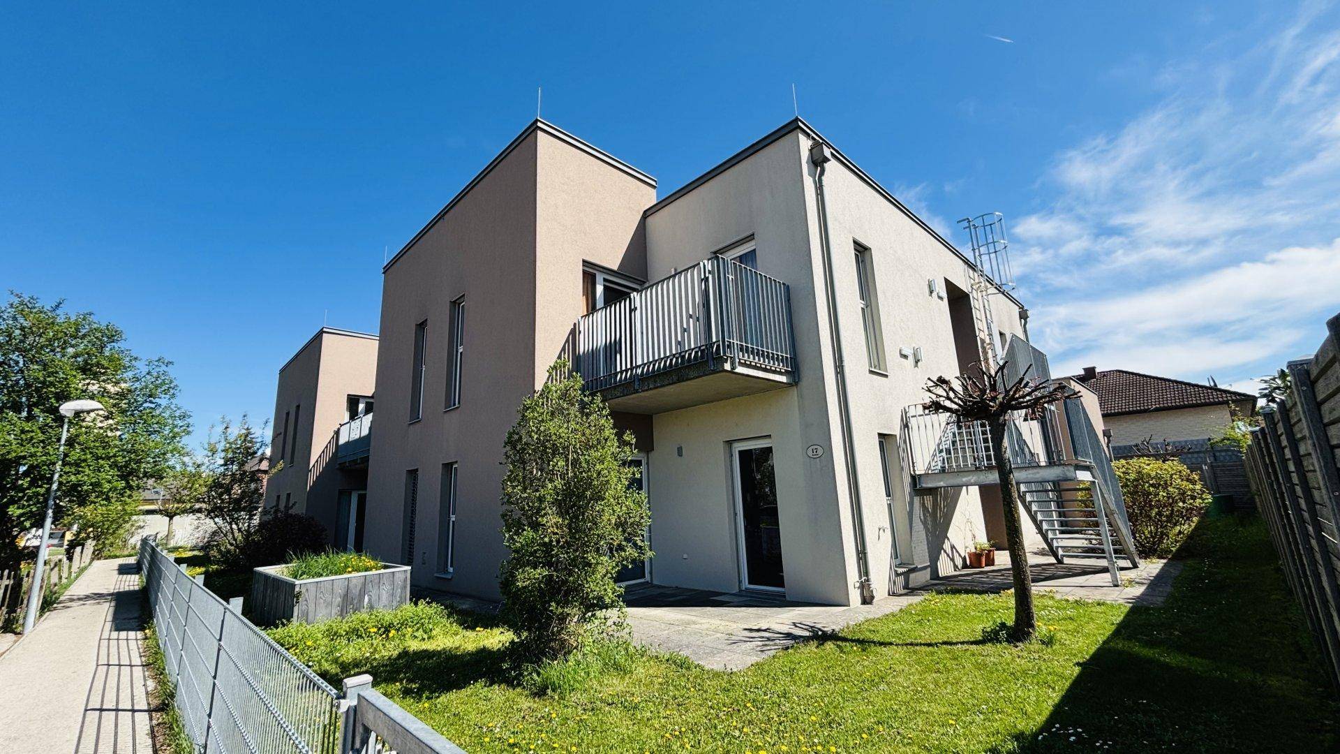 Eckansicht eines Wohnhauses mit Balkon und Zugang zu einer kleinen Grünfläche bei sonnigem Wetter.