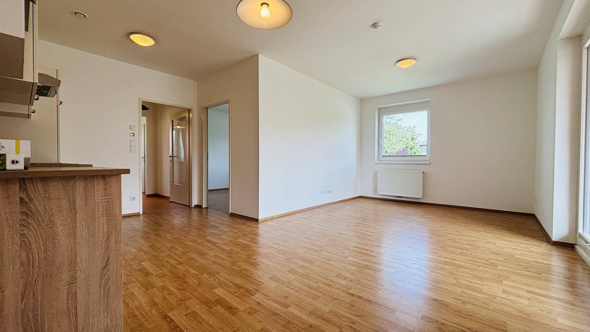 Heller Wohnbereich mit Laminatboden, großem Fenster und offenem Übergang zu weiteren Räumen.