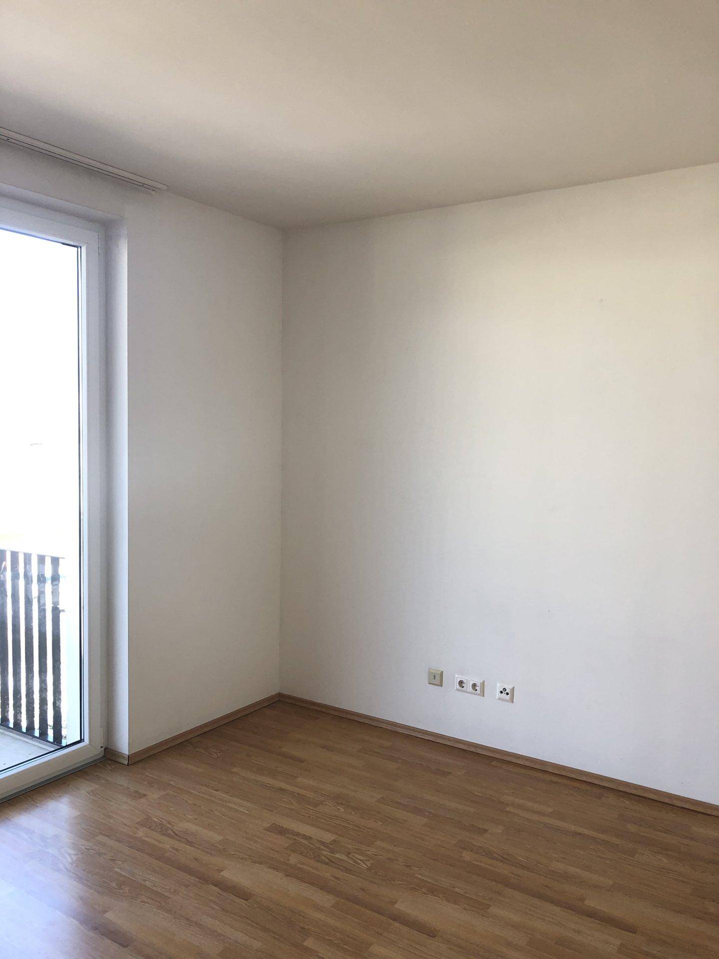 Helles, leeres Zimmer mit Laminatboden und Zugang zu einem Balkon.