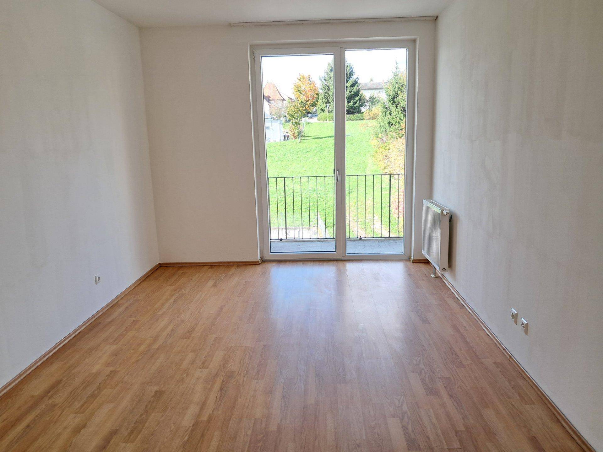 Geräumiges, helles Zimmer mit Laminatboden und Zugang zu einem Balkon mit Grünblick.