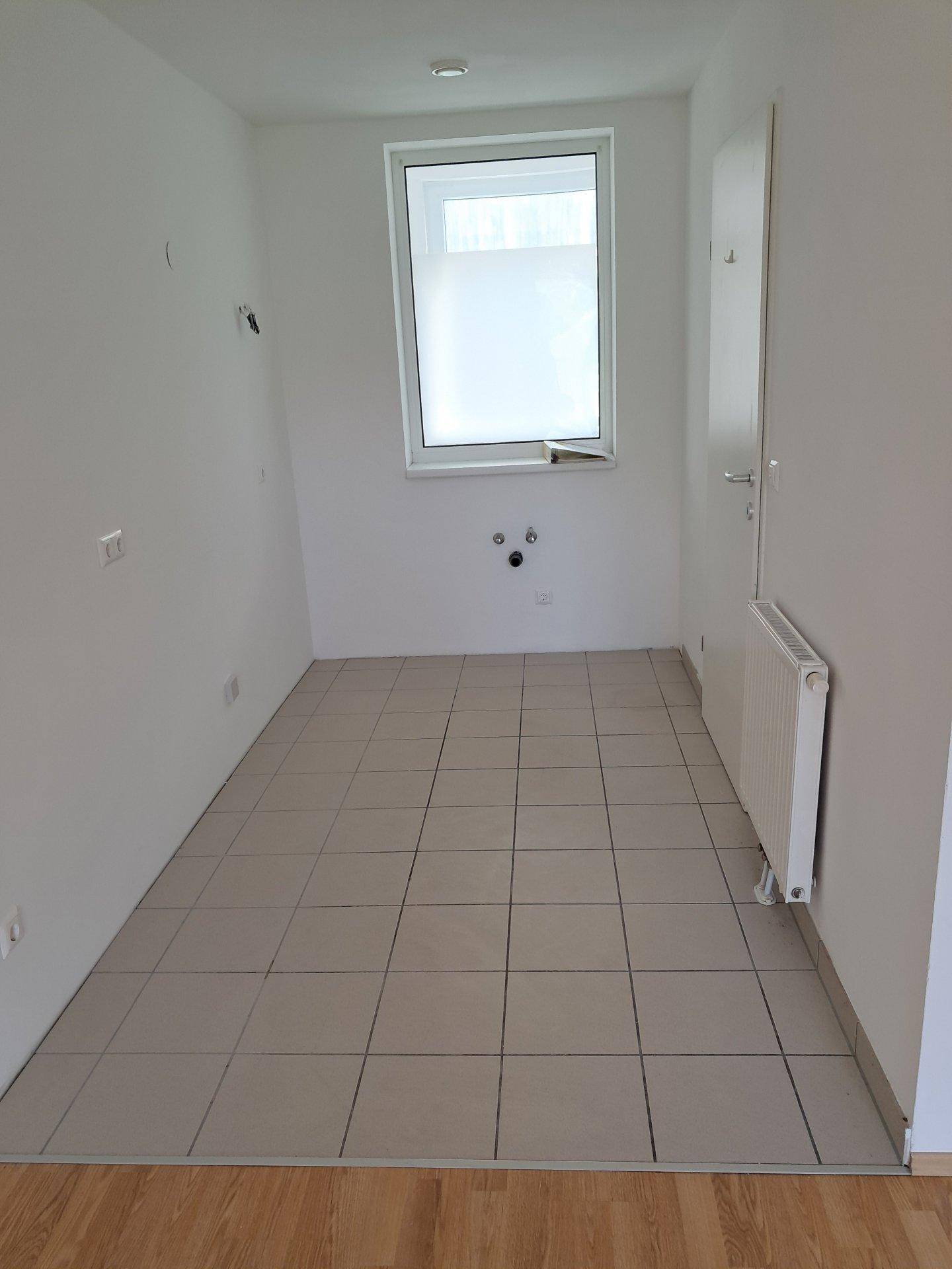 Leerer Raum mit Fliesenboden, Fenster und Anschlüssen für eine Küche oder Waschküche.