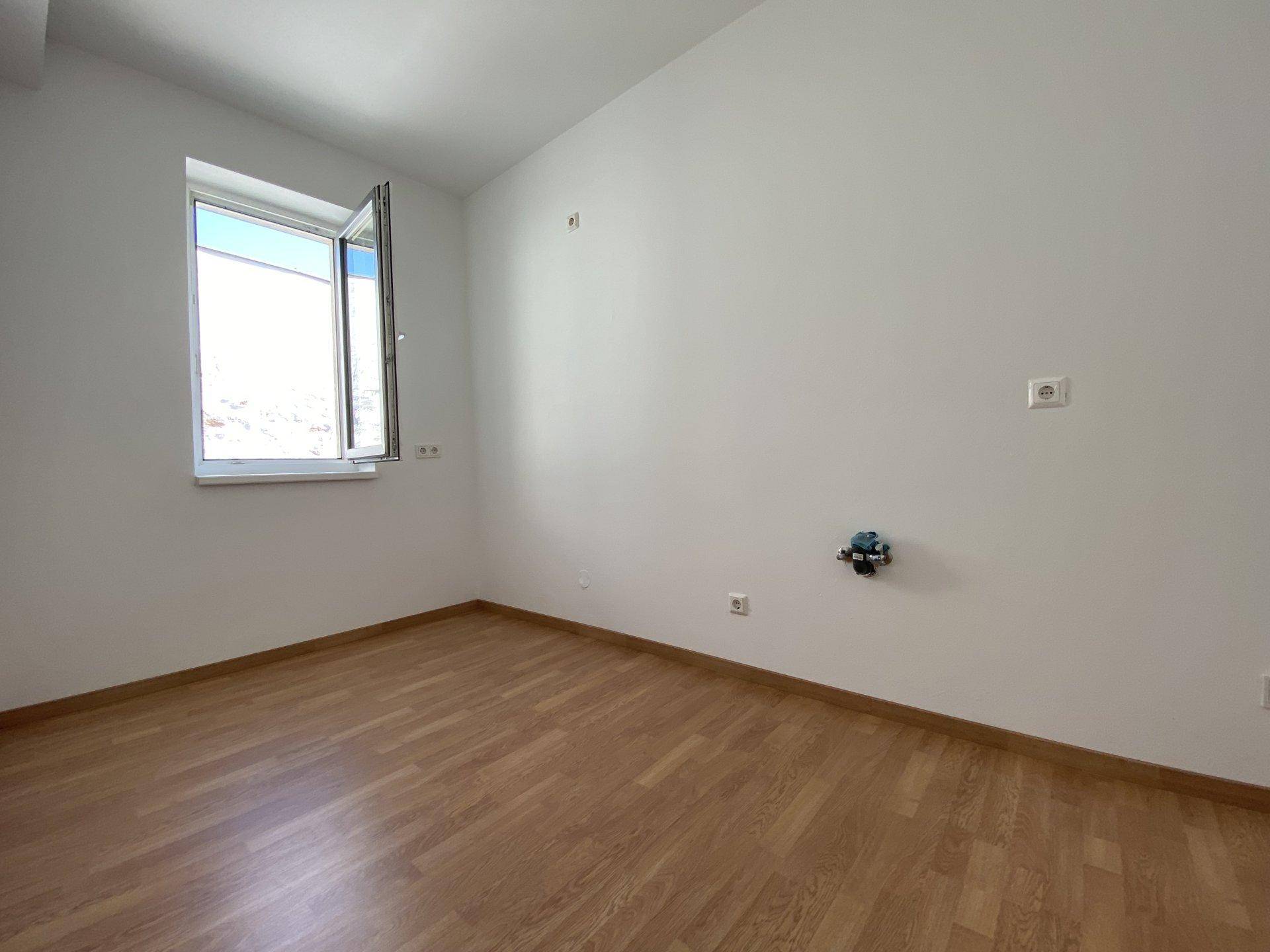 Leeres Zimmer mit Laminatboden und einem geöffneten Fenster, das viel Tageslicht hereinlässt.