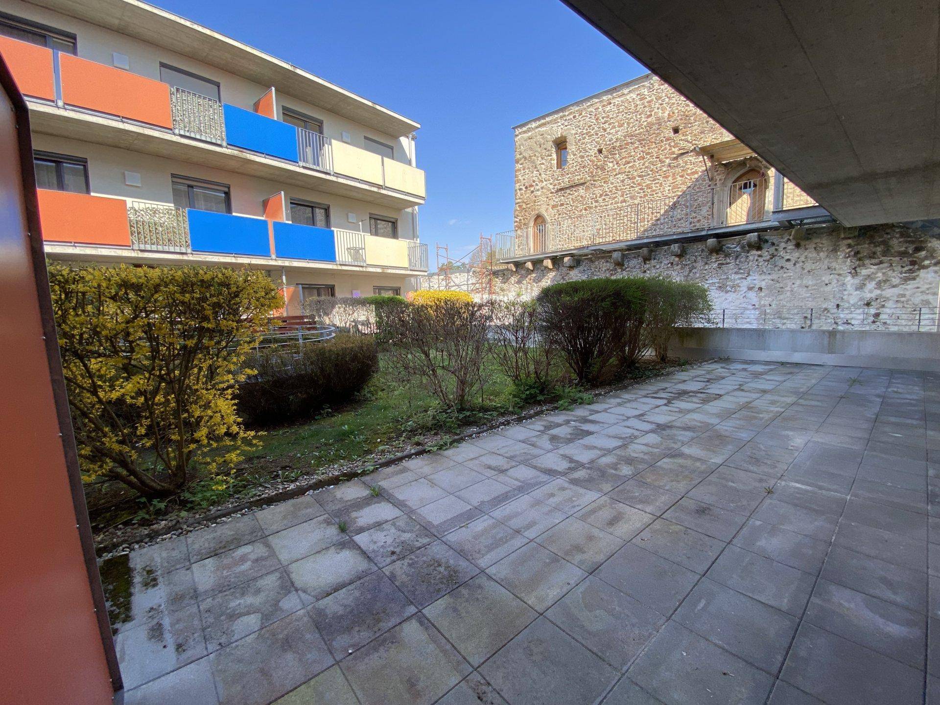 Sonniger Terrassenbereich mit Rasenfläche und einer historischen Steinmauer im Hintergrund.