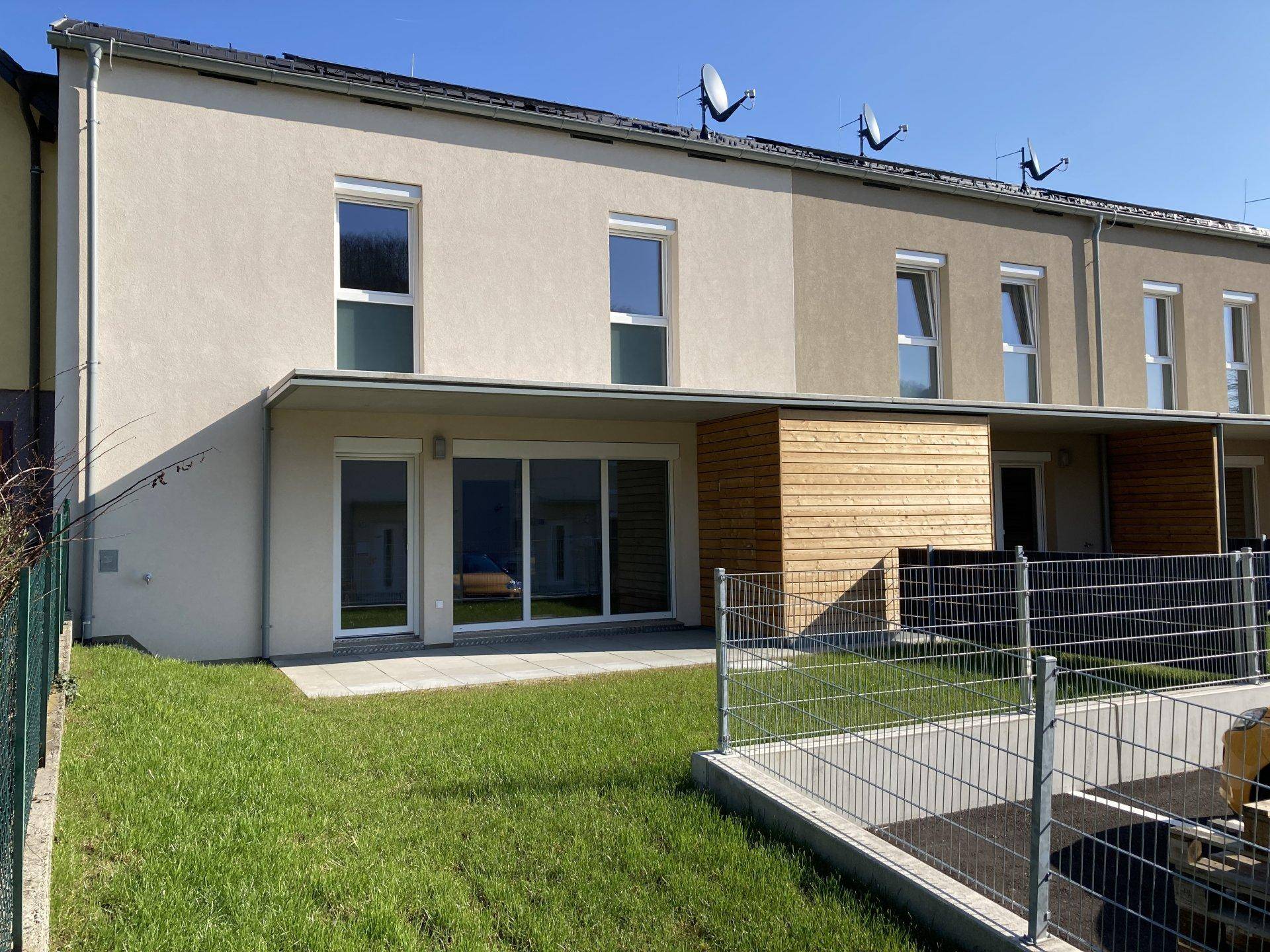 Rückseite eines Reihenhauses mit kleiner Terrasse und Gartenbereich unter blauem Himmel.