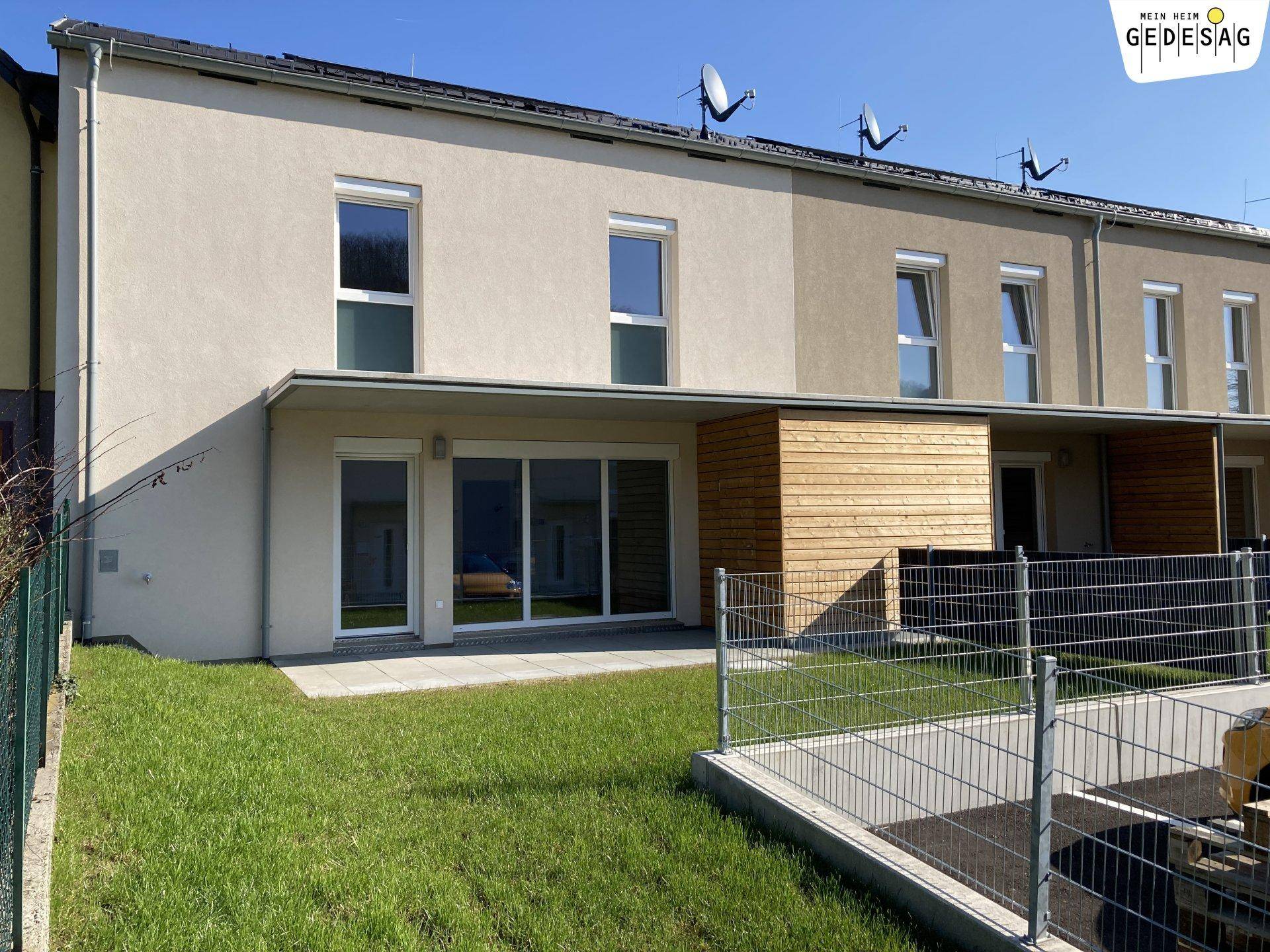 Rückseite eines Reihenhauses mit kleiner Terrasse und Gartenbereich unter blauem Himmel.