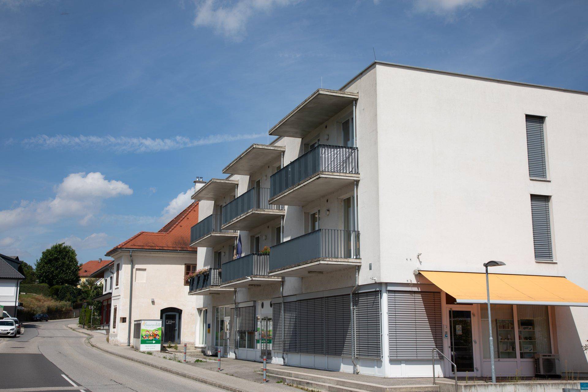 Ansicht des modernen Mehrfamilienhauses mit Balkonen und Geschäften im Erdgeschoss unter blauem Himmel.