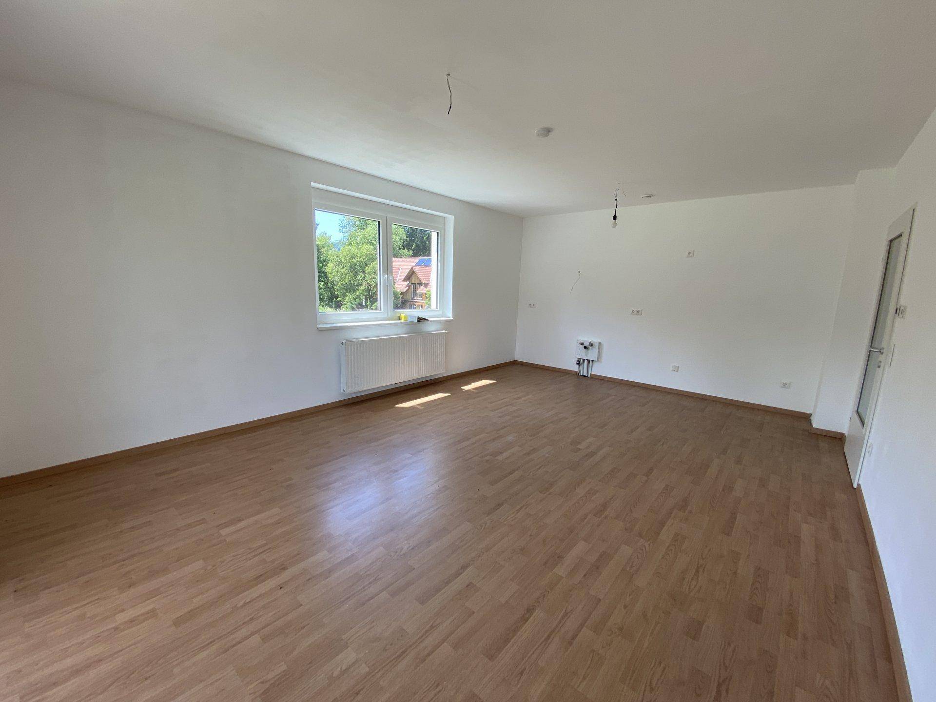Geräumiger, unmöblierter Wohnbereich mit hellem Laminatboden und großen Fenstern mit Blick ins Grüne.