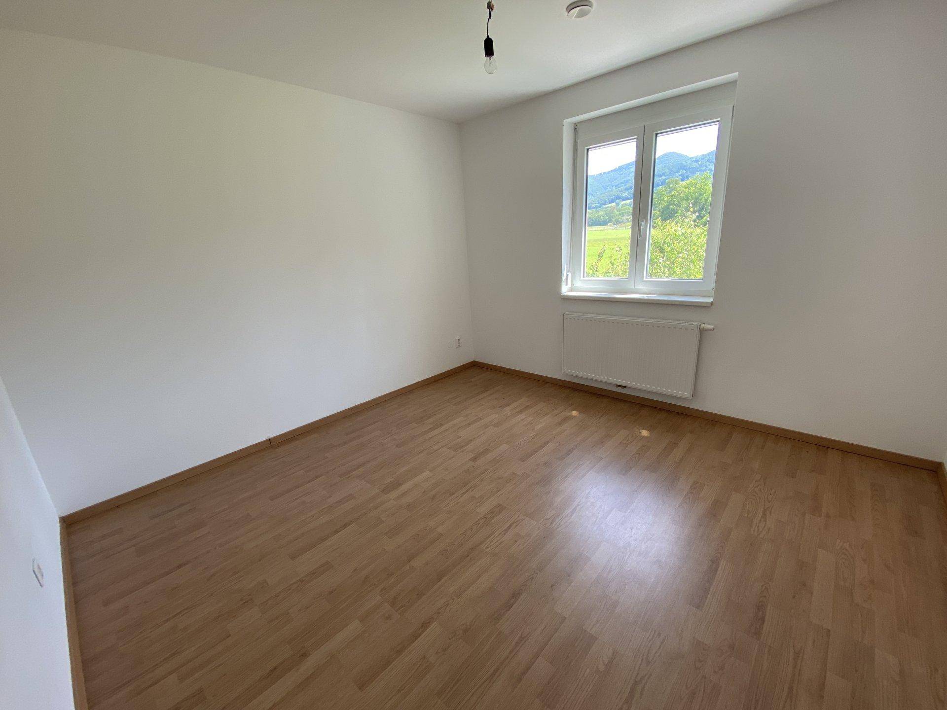 Unmöbliertes Schlafzimmer mit hellem Laminatboden und einem großen Fenster, das viel Tageslicht hereinlässt.