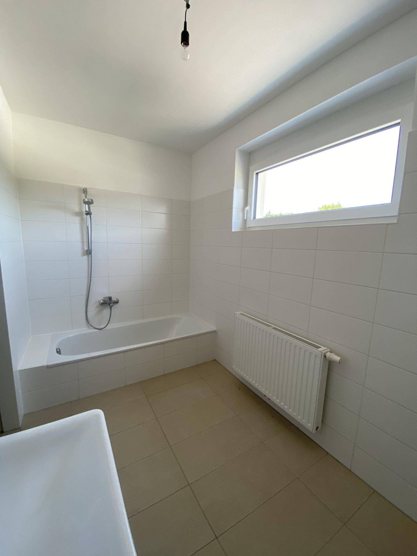 Badezimmer mit weißen Fliesen, Badewanne mit Duschfunktion, Heizkörper und Fenster für Tageslicht.