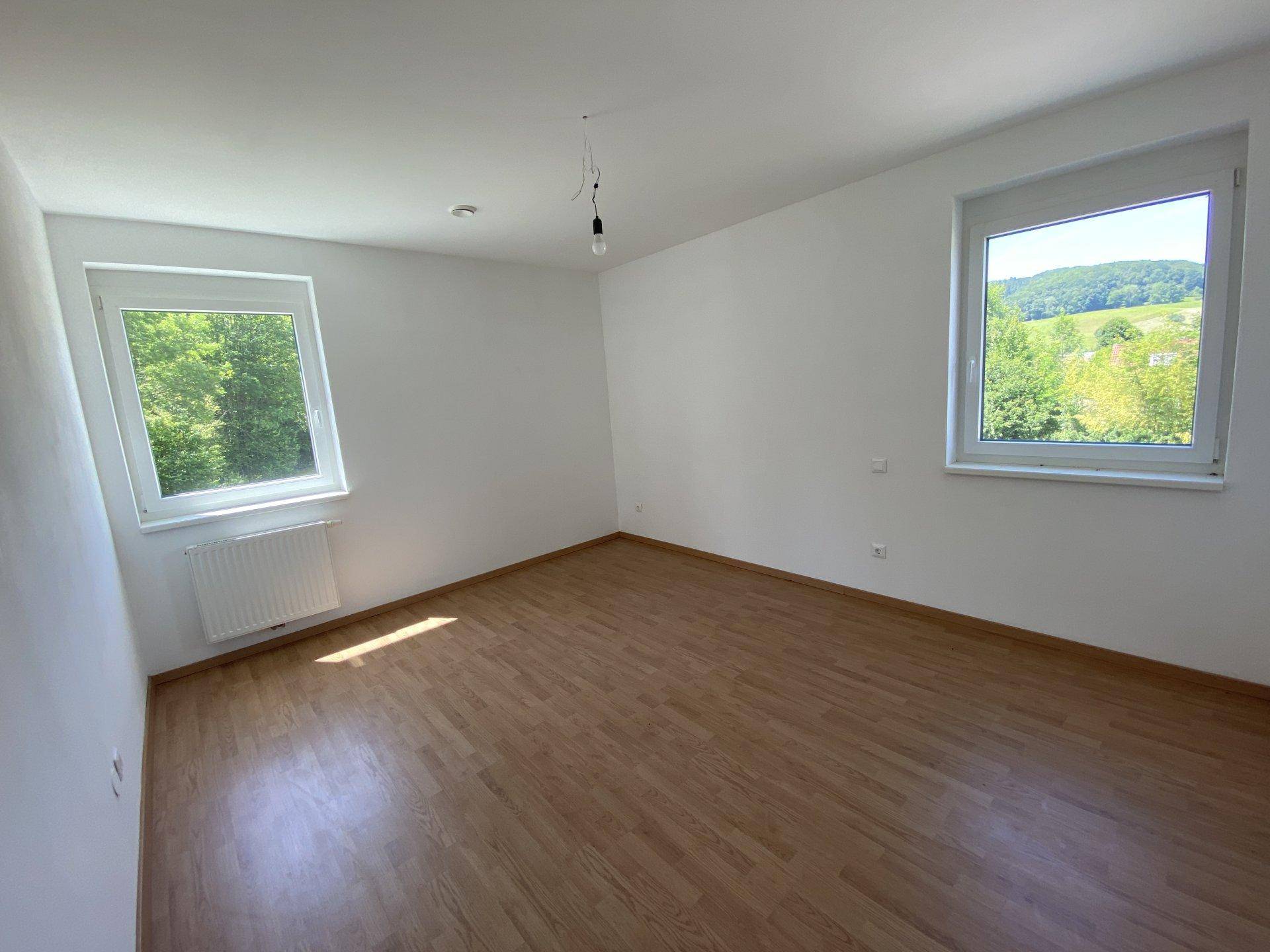 Helles, leeres Schlafzimmer mit Laminatboden, weißen Wänden und zwei Fenstern mit Blick ins Grüne.