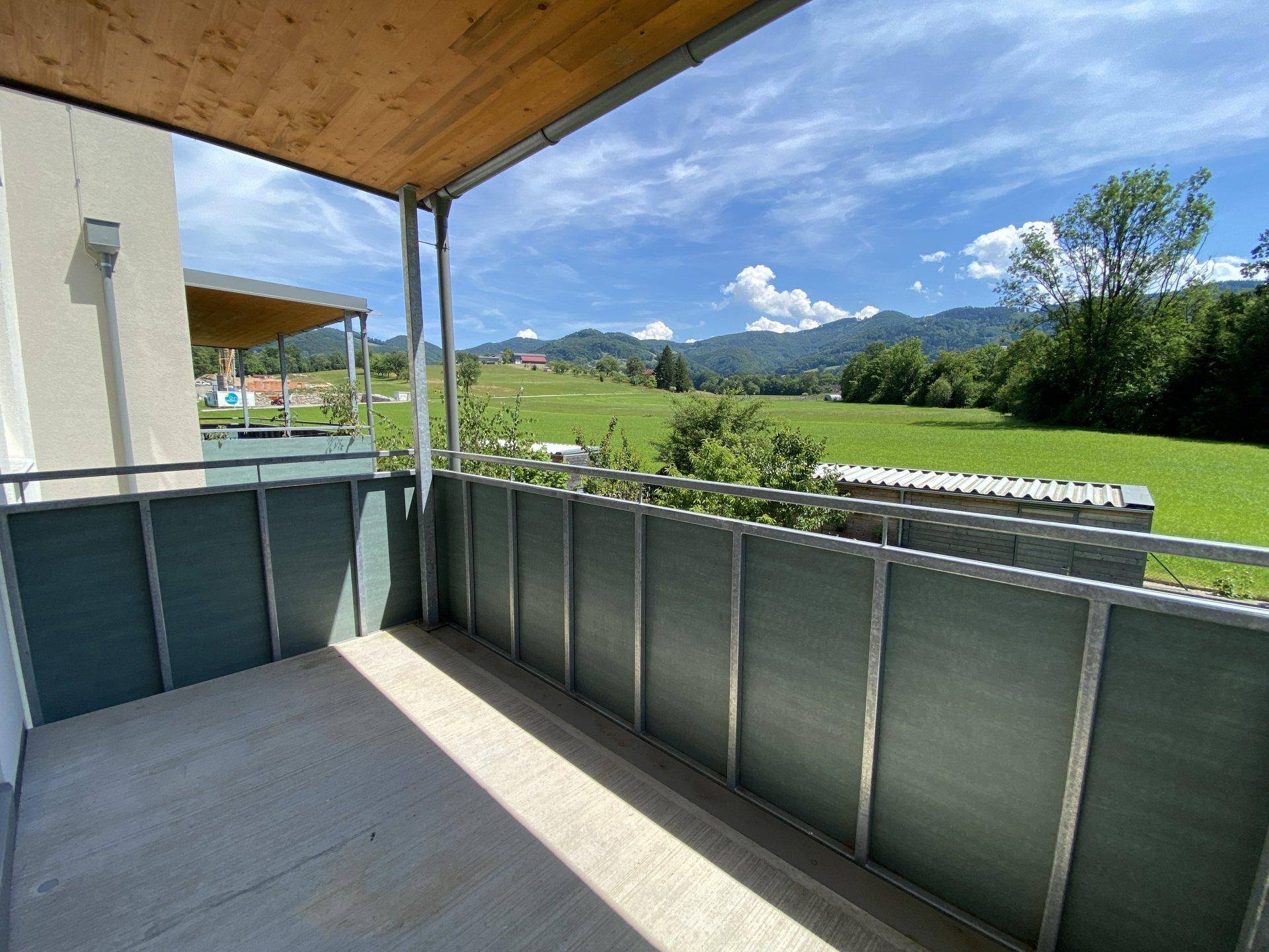 Balkon mit weitem Blick über grüne Felder und sanfte Hügel unter blauem Himmel.