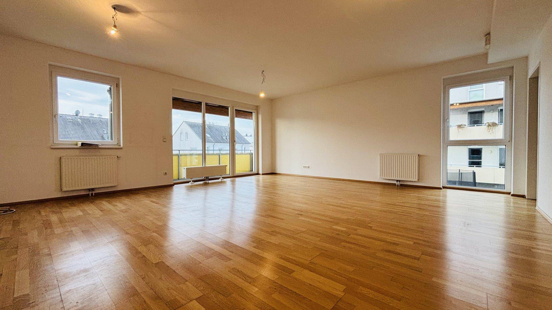 Lichtdurchflutetes Wohnzimmer mit glänzendem Parkettboden und großen Fenstern, die viel Tageslicht hereinlassen.