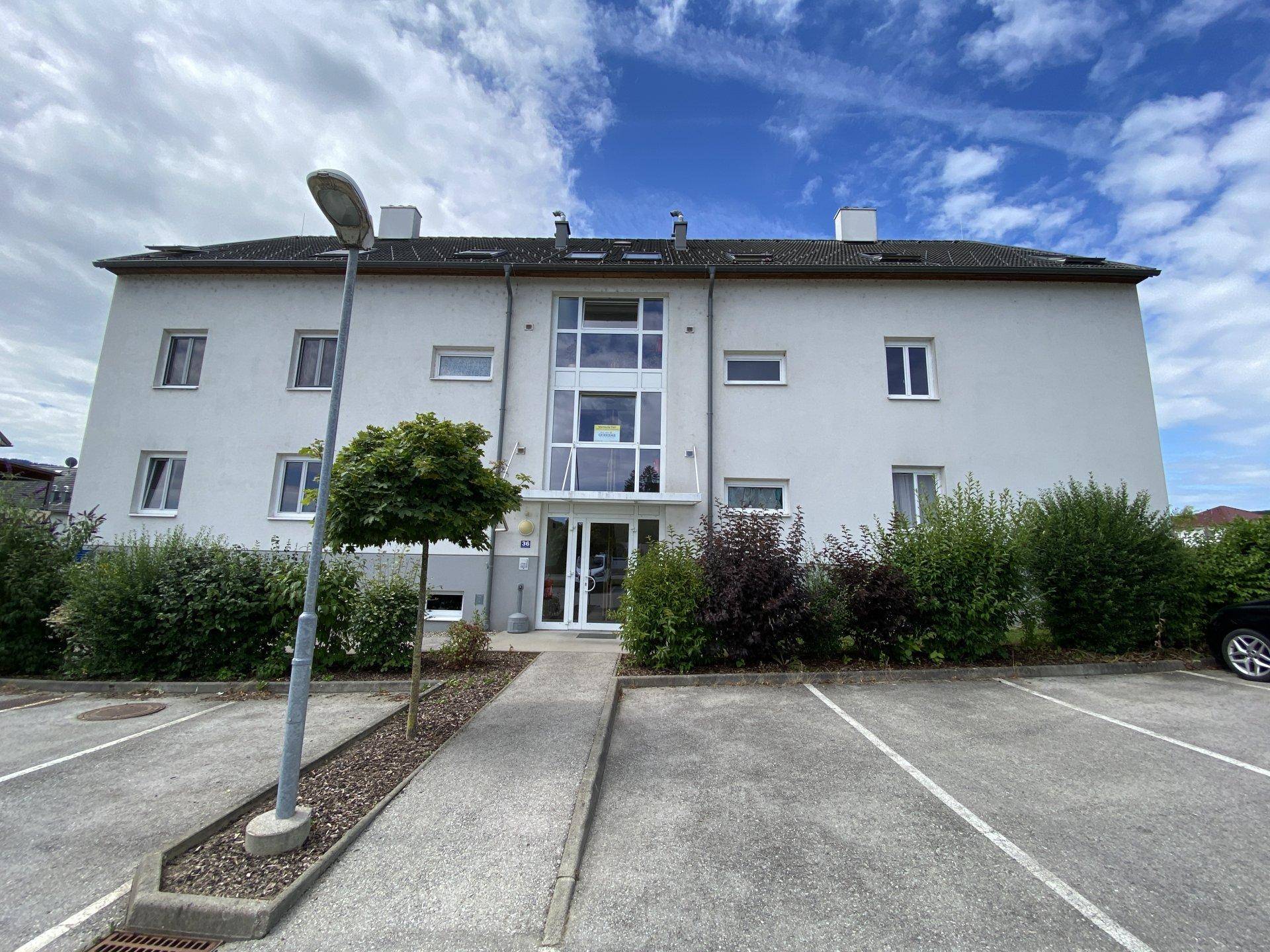 Moderne Wohnhausfassade mit großen Fenstern und Eingangsbereich, umgeben von Grünflächen und Parkplätzen.