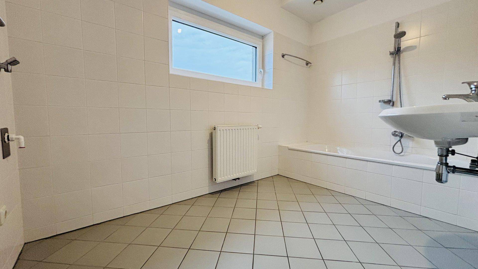 Funktionales Badezimmer mit weißen Fliesen, Badewanne und Waschbecken, ausgestattet mit einem Fenster.