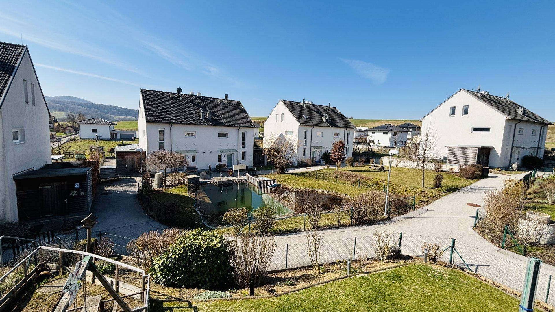 Weitläufige Wohnanlage mit mehreren Häusern, Gärten und einem Teich unter strahlend blauem Himmel.