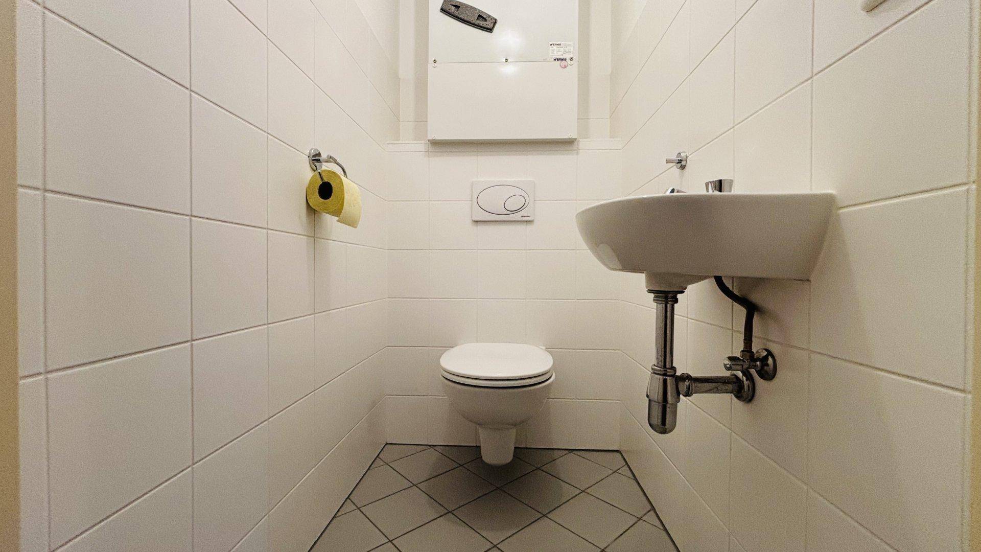 Kleines Gäste-WC mit weißen Fliesen, wandhängendem WC und Handwaschbecken, praktisch und sauber.