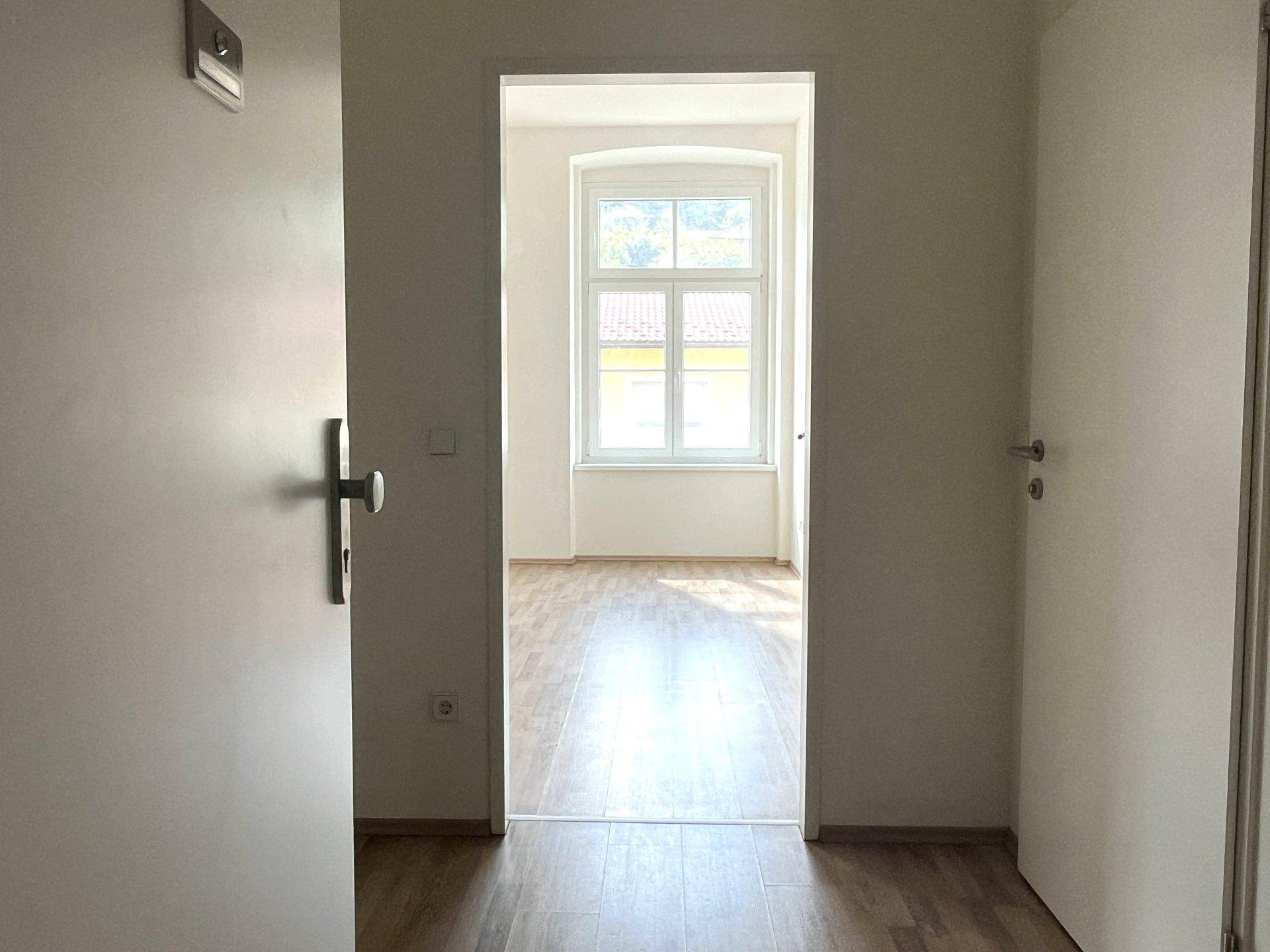 Blick durch eine Tür in einen hellen Raum mit Laminatboden und großem Fenster, das Tageslicht hereinlässt.