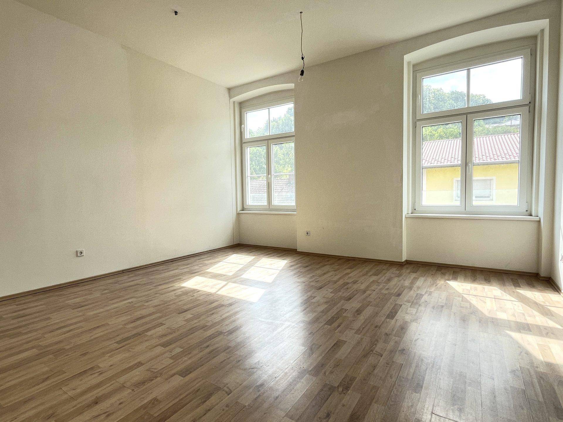 Heller, leerer Raum mit Laminatboden und zwei großen Fenstern, die viel Sonnenlicht hereinlassen.