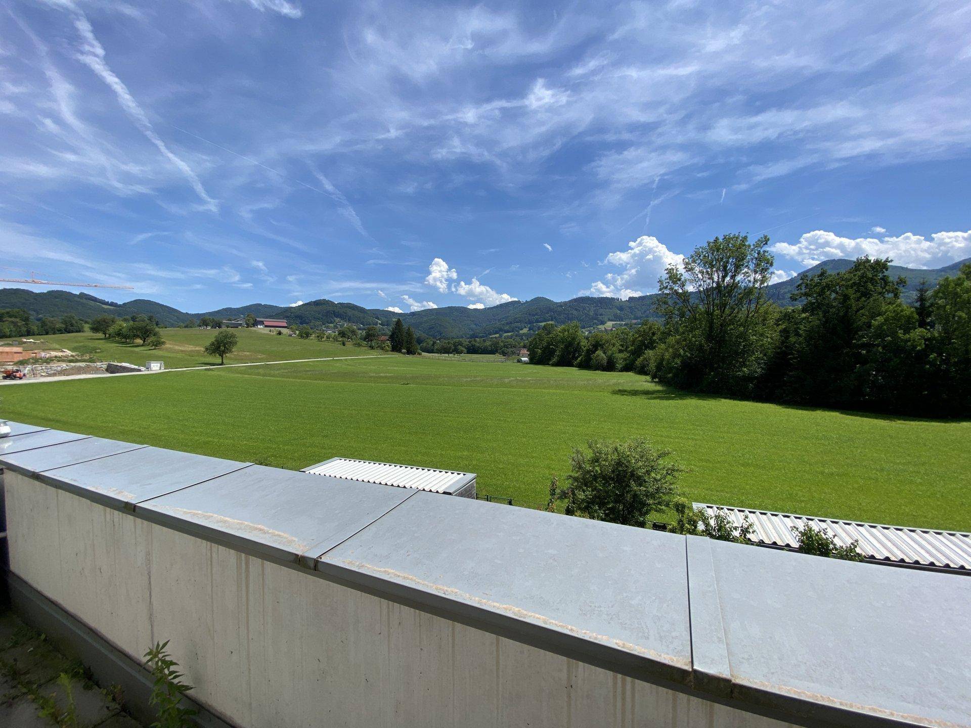 Panoramablick von der Terrasse auf die weite grüne Landschaft mit Hügeln und Bäumen unter blauem Himmel.
