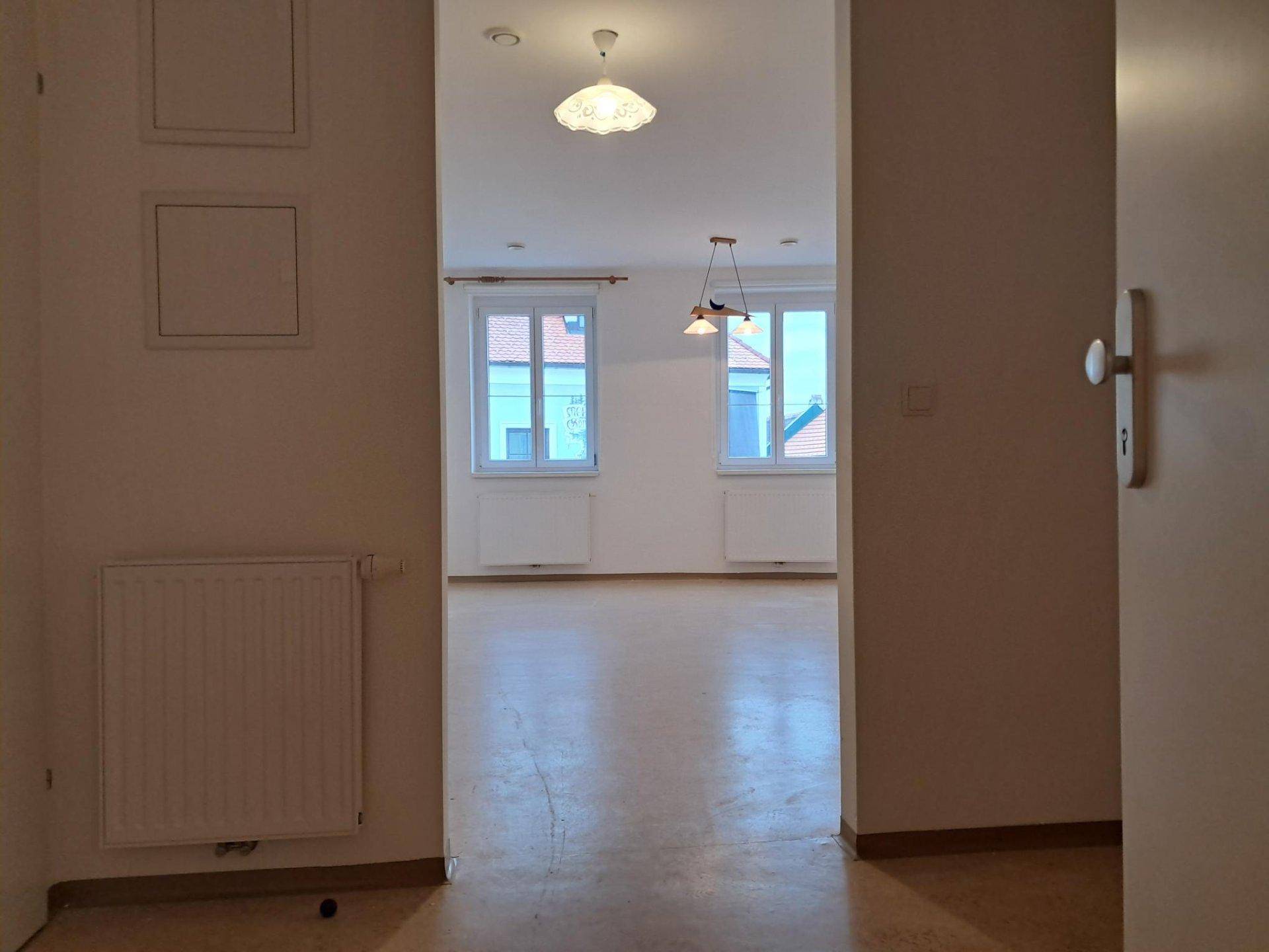 Blick vom Flur in einen hellen, leeren Raum mit zwei Fenstern und Heizkörpern.
